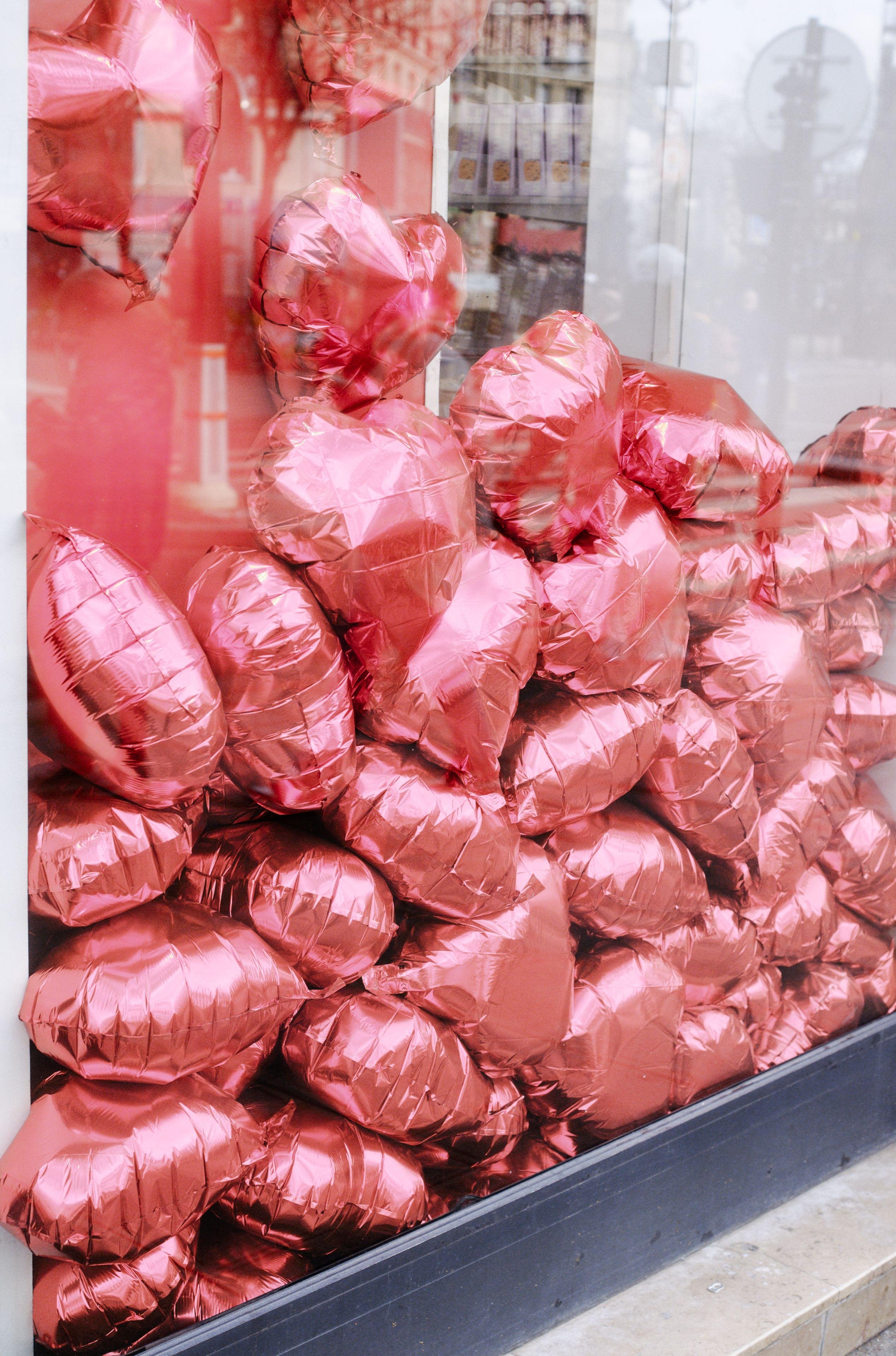 Photo verticale. Derrière une vitrine, de nombreux ballons en forme de cœur sont entassés contre la vitre.
