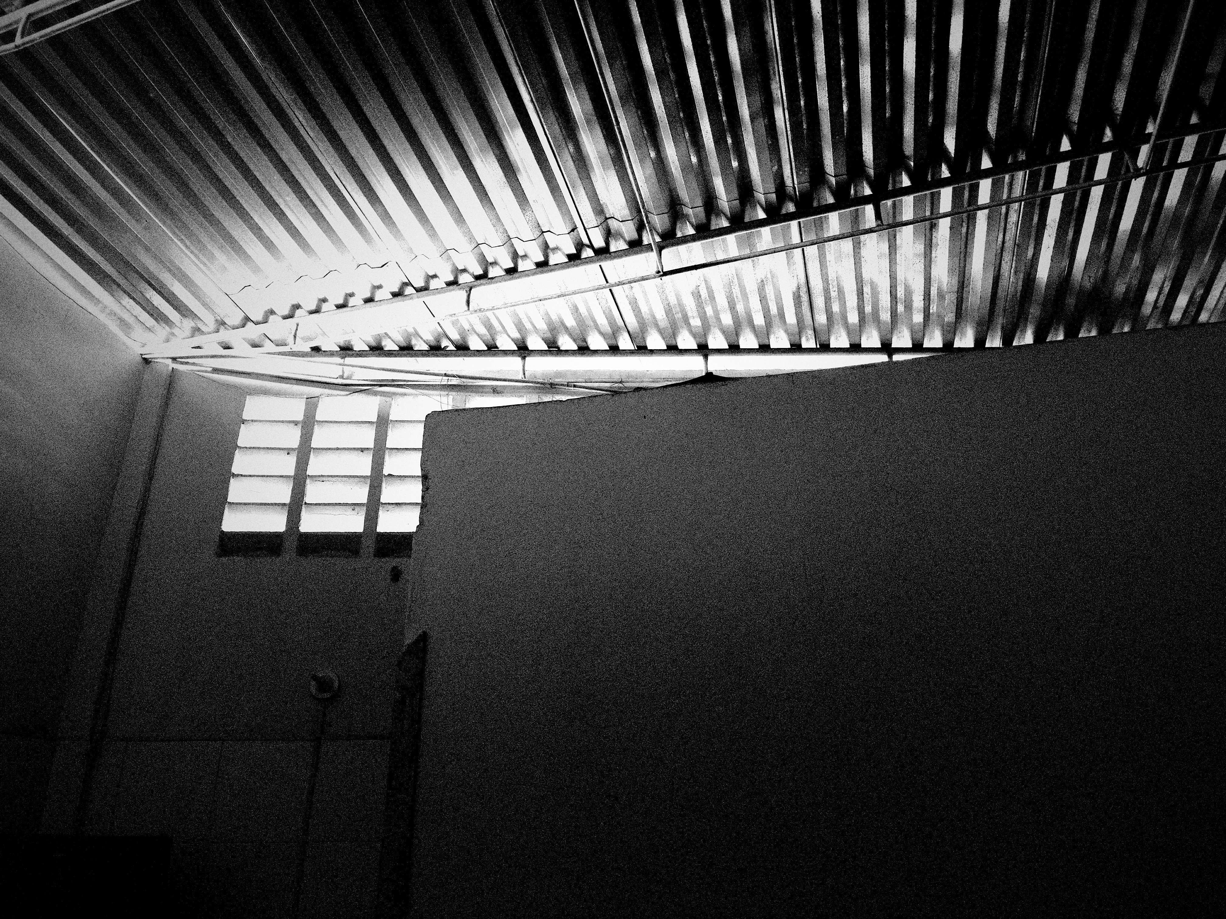 Corrugated metal ceiling with light streaming through, creating high contrast shadows. A partial wall divides the space below. Vertical slatted windows allow additional light in. The scene is monochromatic, emphasizing patterns and angles. 