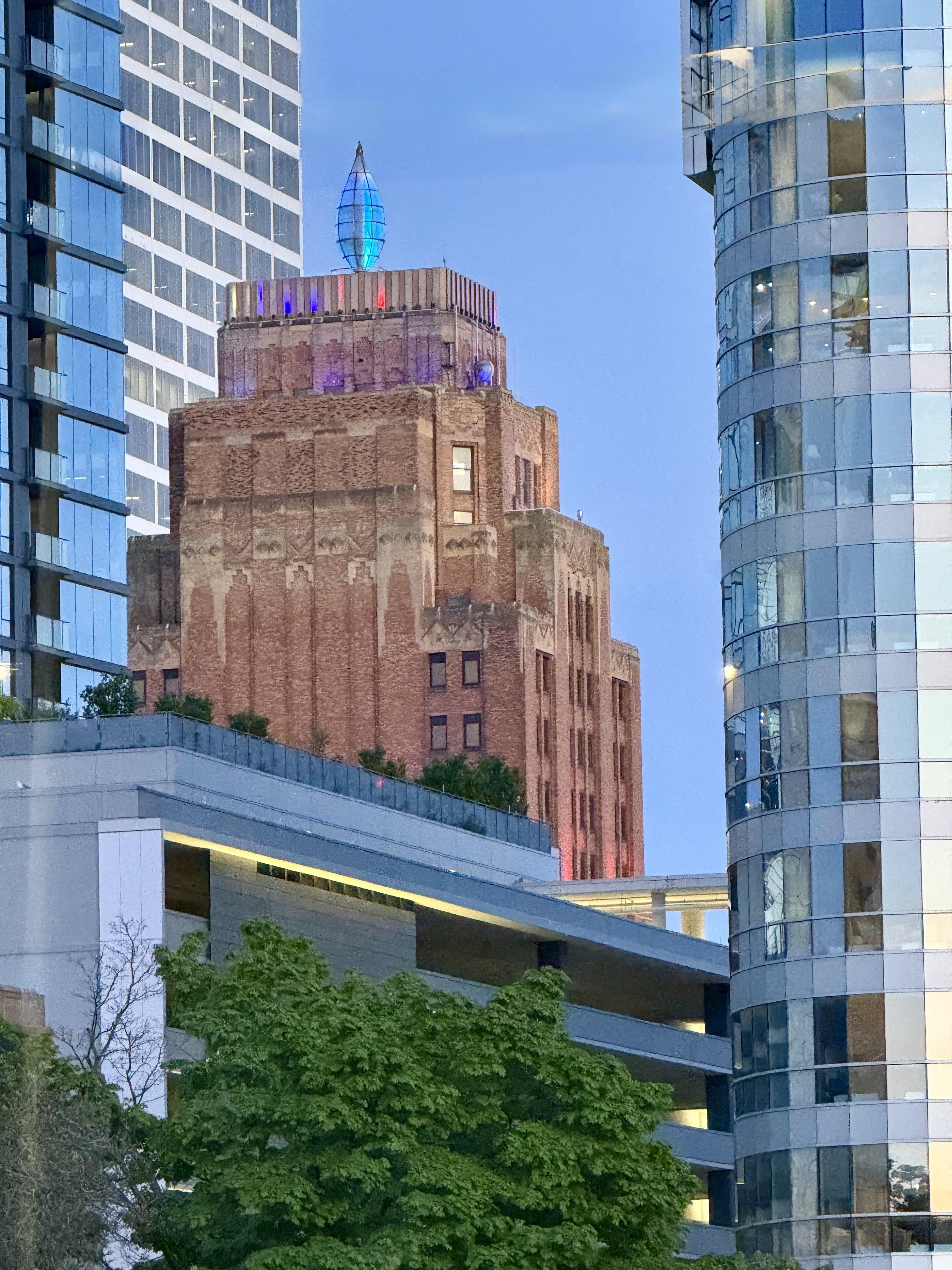 Red brick building with an ornate top and a blue, glass-like spire. Surrounded by modern buildings with reflective glass, some with curved facades. A foreground of green trees partially obscures the lower structures. The sky is clear and blue.