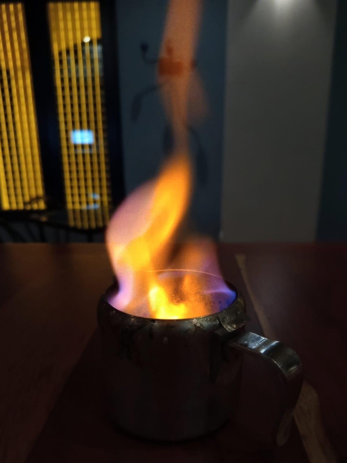 Una barra de bar de madera con una jarra metálica con alcohol quemándose para hacer un asiático. Sale una llama de fuego grande de colores naranjas, rosas y azules