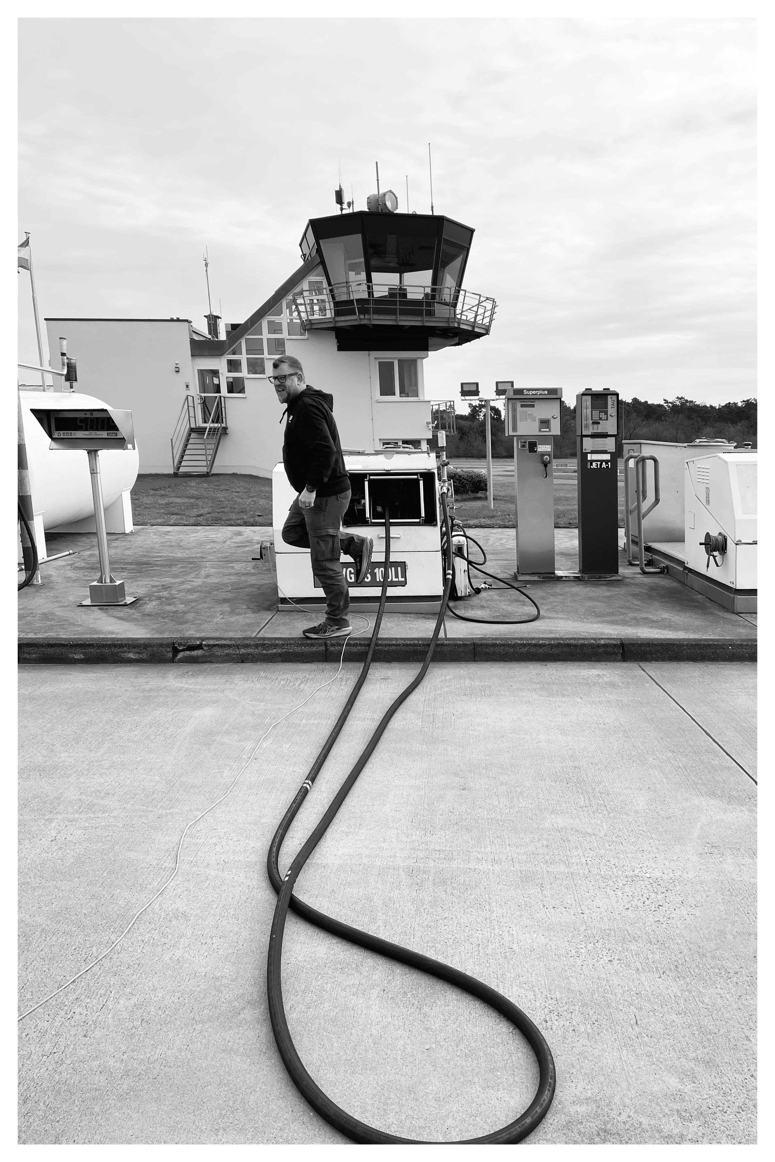 Man standing beside a fuel pump at a small airfield, with two long hoses snaking across the concrete foreground toward the pump; a control-tower building and additional fueling equipment are in the background.