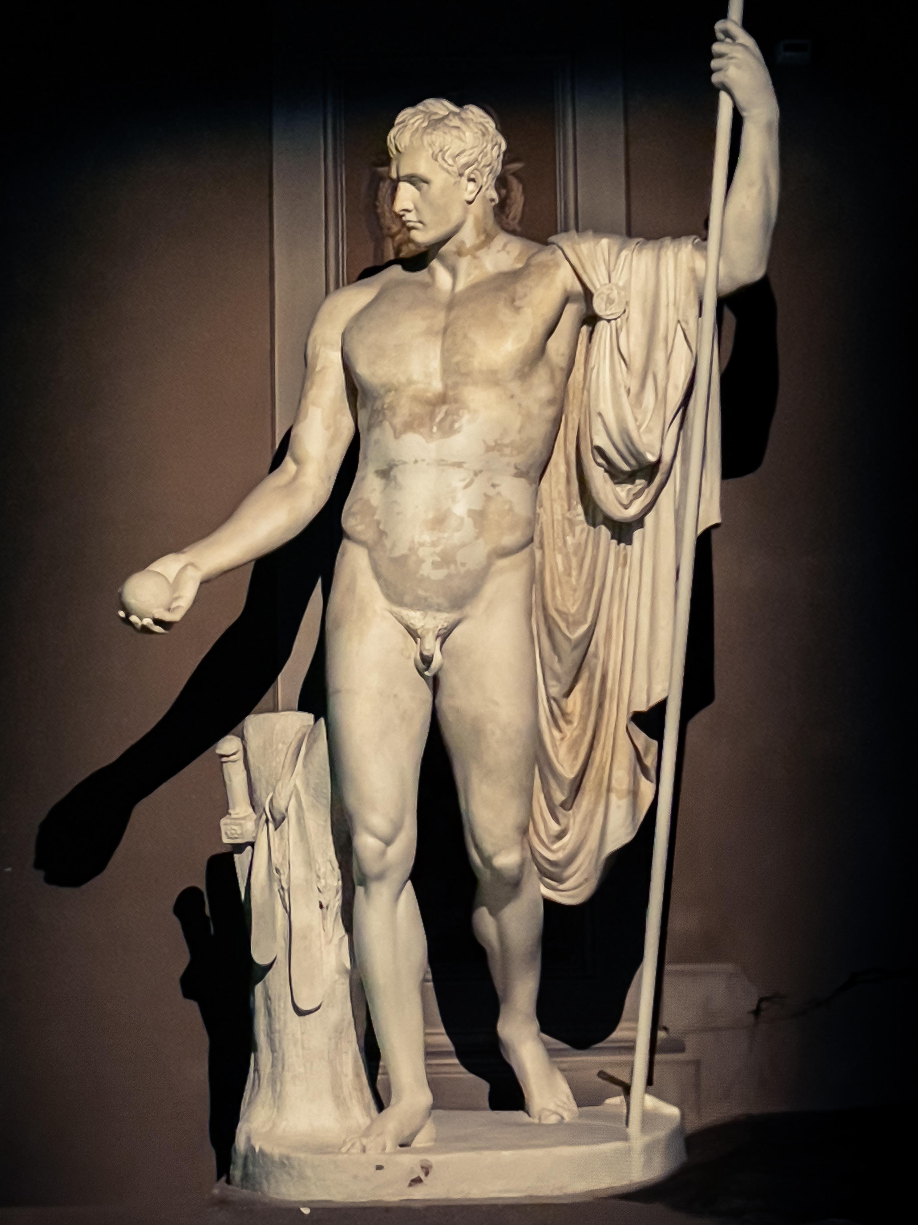 Statua classica in marmo raffigurante un giovane uomo con mantello drappeggiato, lancia in mano e un oggetto nella mano sinistra, posizionata in un ambiente museale con illuminazione che evidenzia i dettagli scolpiti.