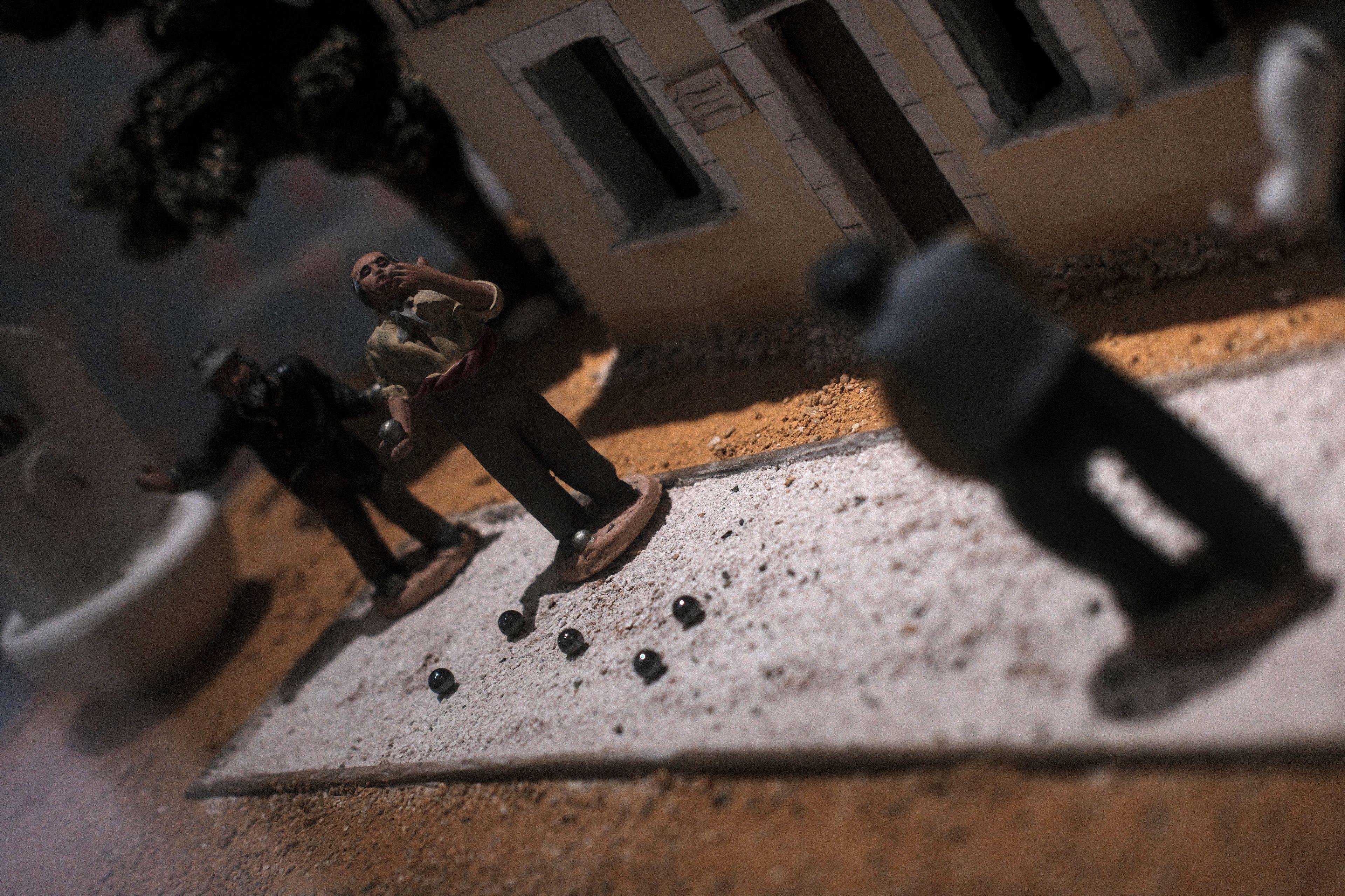 Photo horizontale très penchée. Un diorama fait en santons (des figurines en argile cuite et peinte) représentant une partie de pétanque avec 2 personnages côté résultat et 2, flous, côté lancé