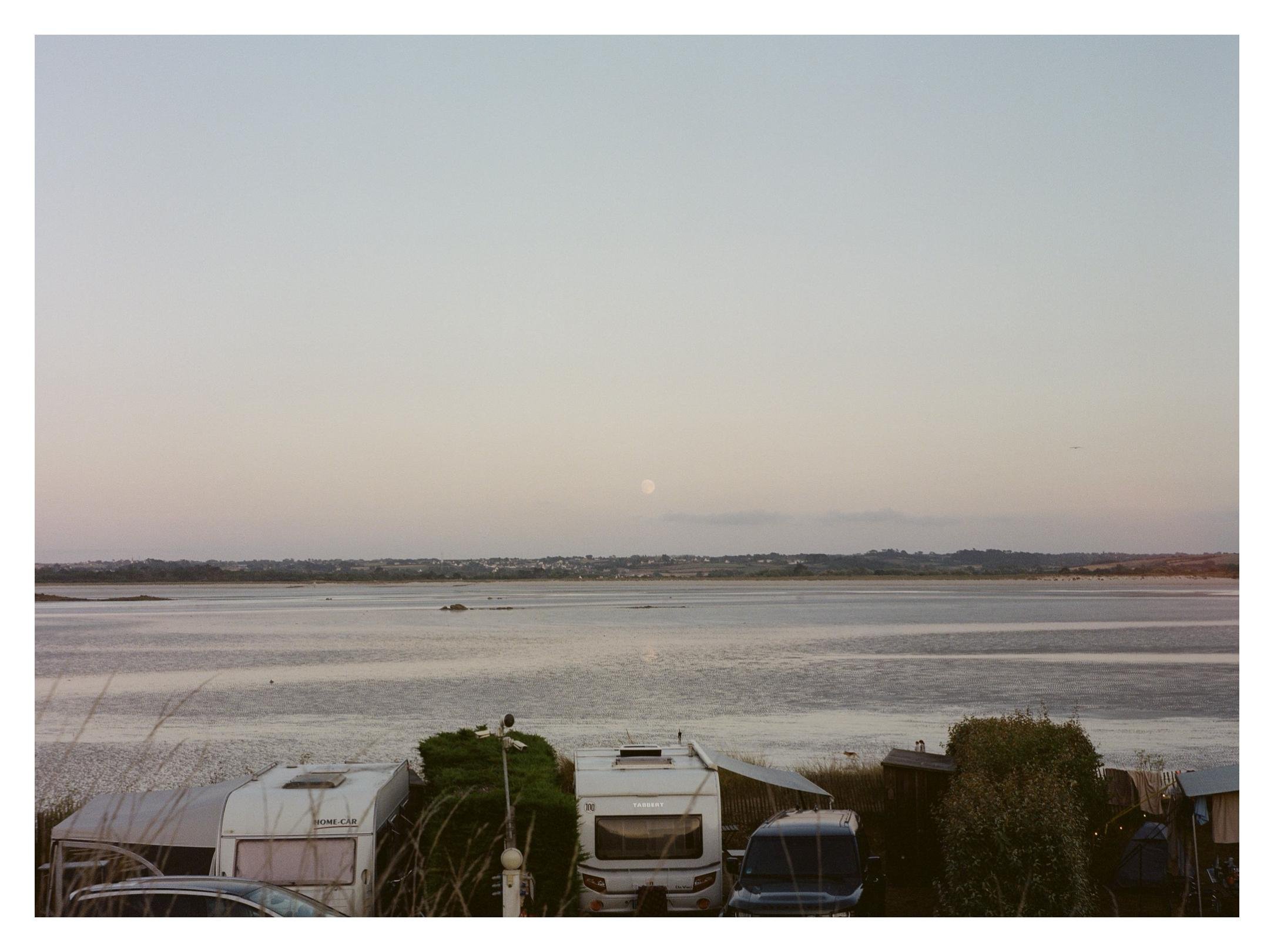Wide view of a calm bay at dusk with a pale moon above the distant shoreline; in the foreground, several parked caravans and vehicles with shrubs partially obscuring them.