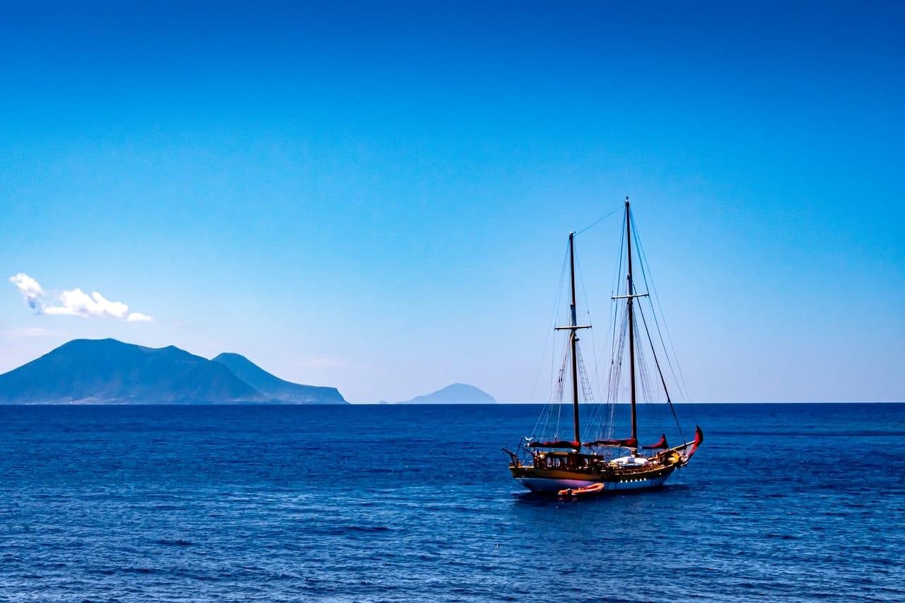Two-masted sailboat on calm blue ocean, positioned right of center, with dark islands on the left horizon under a clear blue sky.