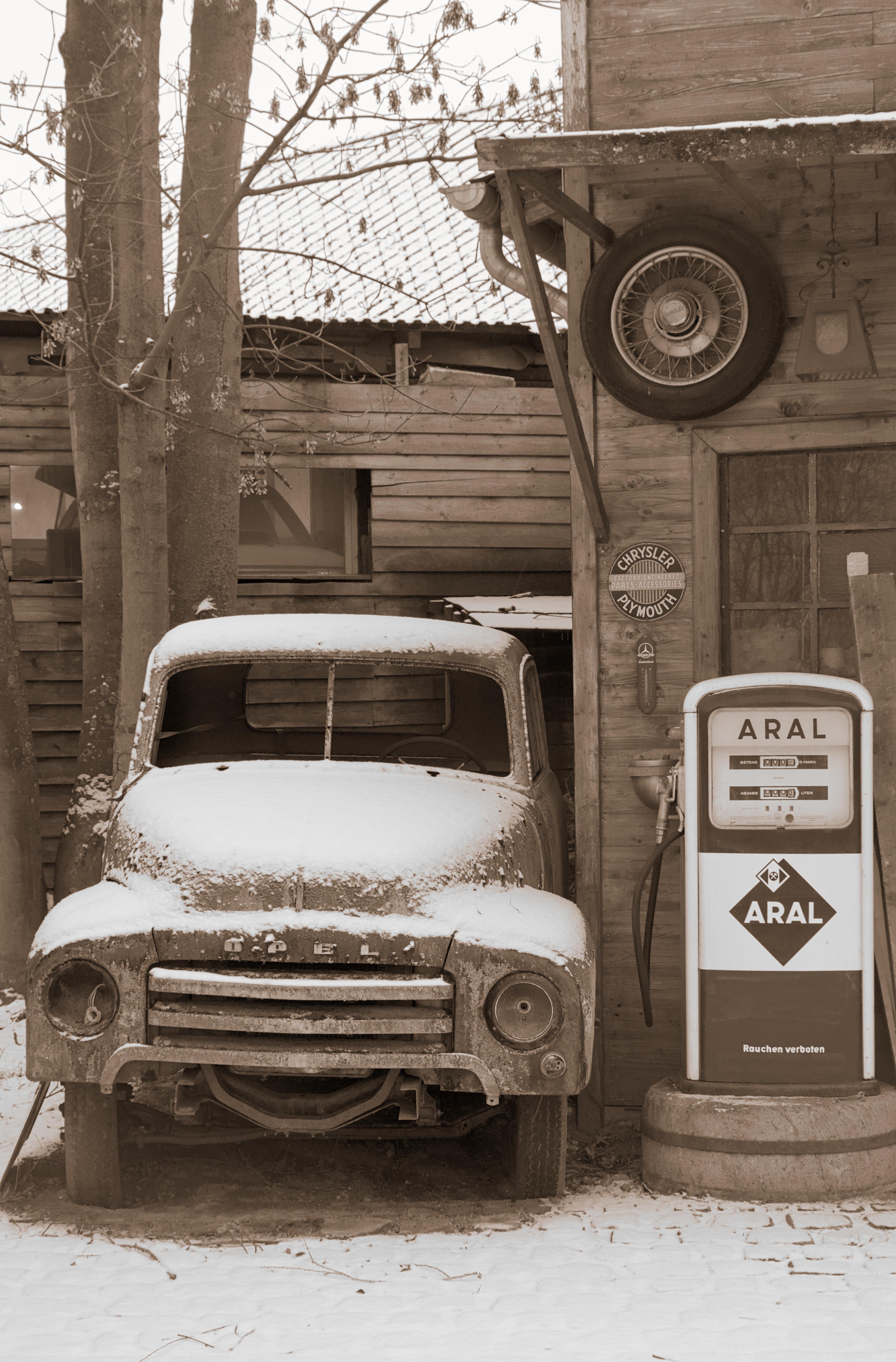 The image depicts a vintage scene with an old, snow-covered Opel truck parked beside a wooden building. There is an old ARAL fuel pump next to the truck, and a spare tire is mounted on the wall above. The overall atmosphere has a nostalgic, rustic feel, possibly set in a snowy rural area.