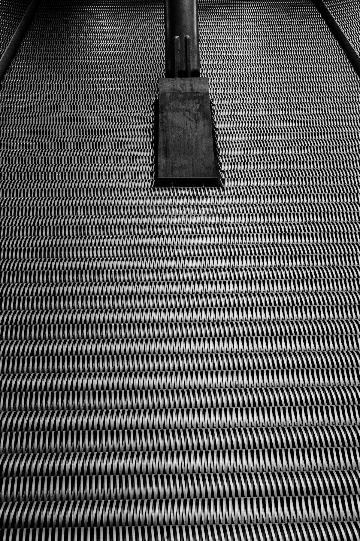 Photo verticale en noir et blanc. Contre plongée sur un mur métallique texturé de traits de travers, une ligne sur deux, vers la droite et vers la gauche. Au sommet de l’image, de début d’une poutre sortant du mur