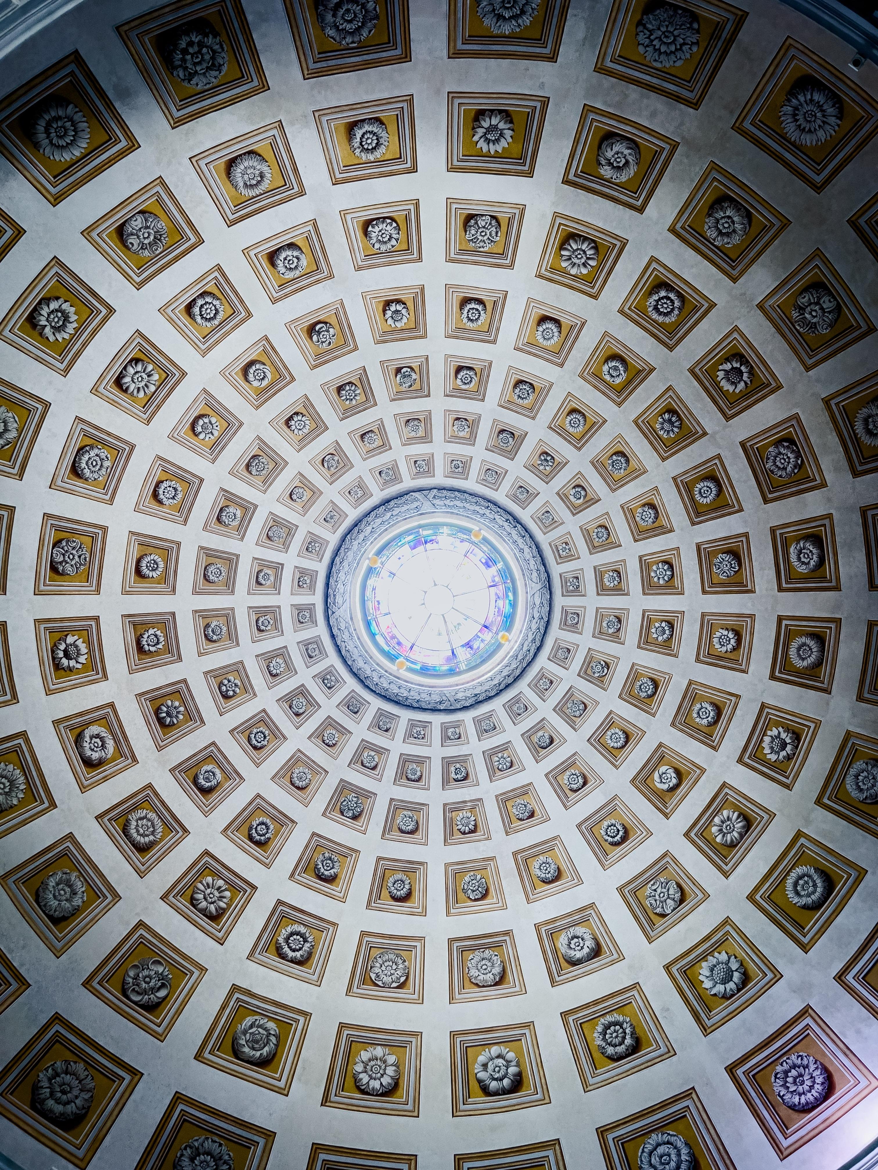 Soffitto di una cupola, progettato con intricati motivi geometrici. La cupola è divisa in numerosi pannelli quadrati, ognuno dei quali presenta dettagliati motivi floreali. Al centro, c'è una finestra circolare di vetro colorato che permette alla luce di entrare, creando un punto focale per il design.