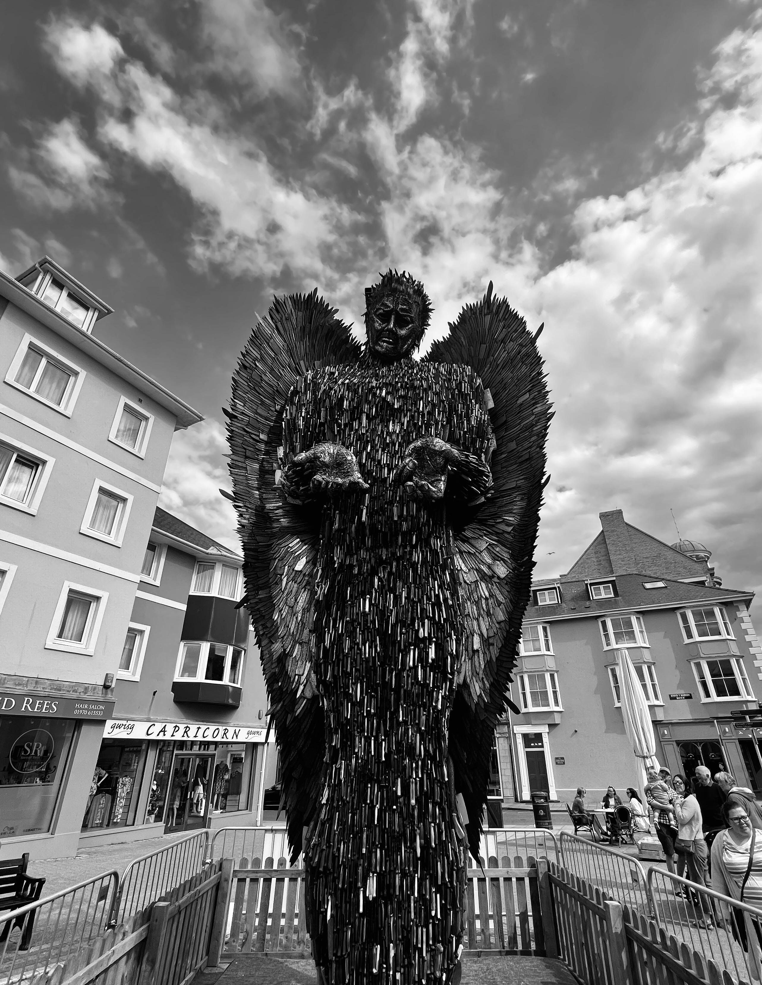 Tall winged statue made of knives, standing behind a small fenced barrier in a town square, with buildings and shopfronts on both sides, a few pedestrians nearby, and a cloudy sky overhead.