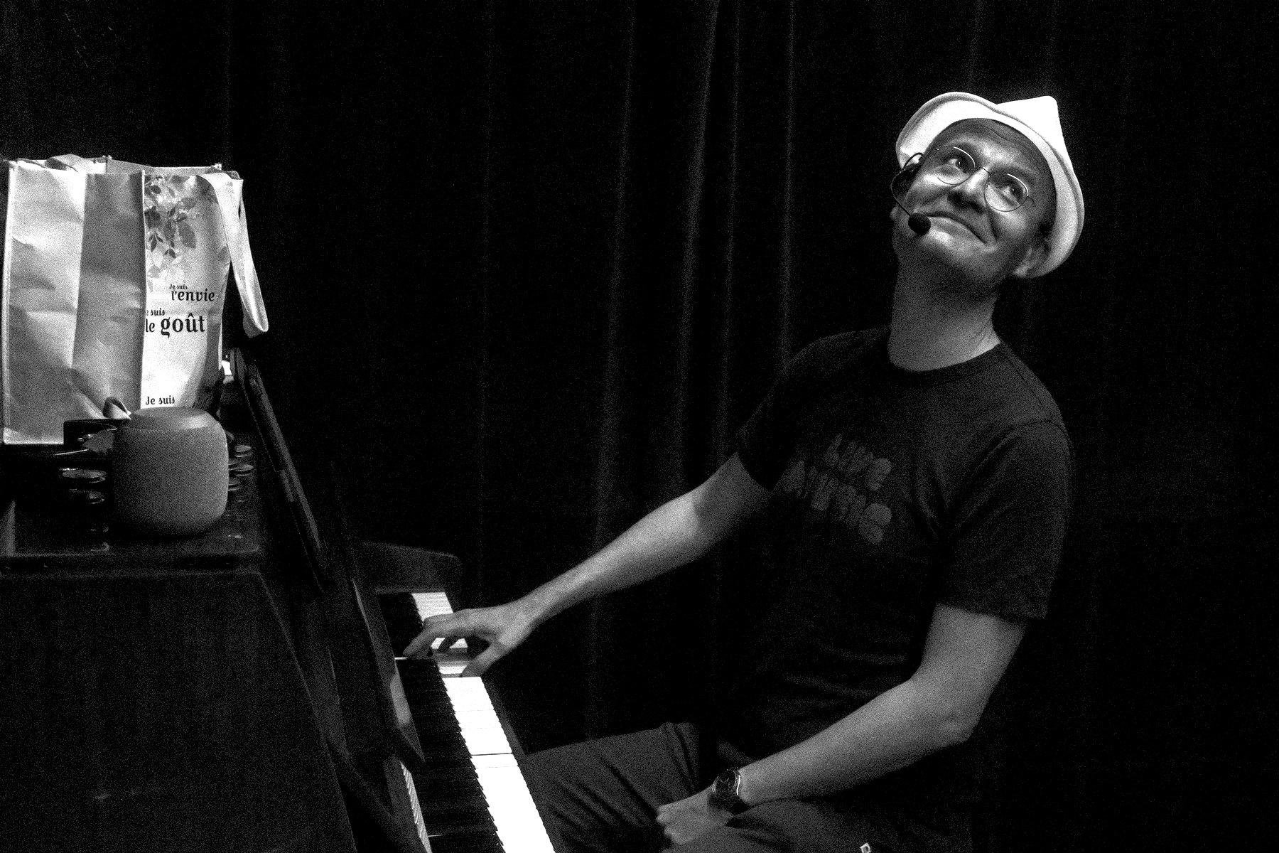 Photo horizontale en noir et blanc. Un homme au chapeau blanc a sa main droite sur un piano et penche sa tête en arrière pour regarder son comparse avec un sourire mi moqueur mi amusé