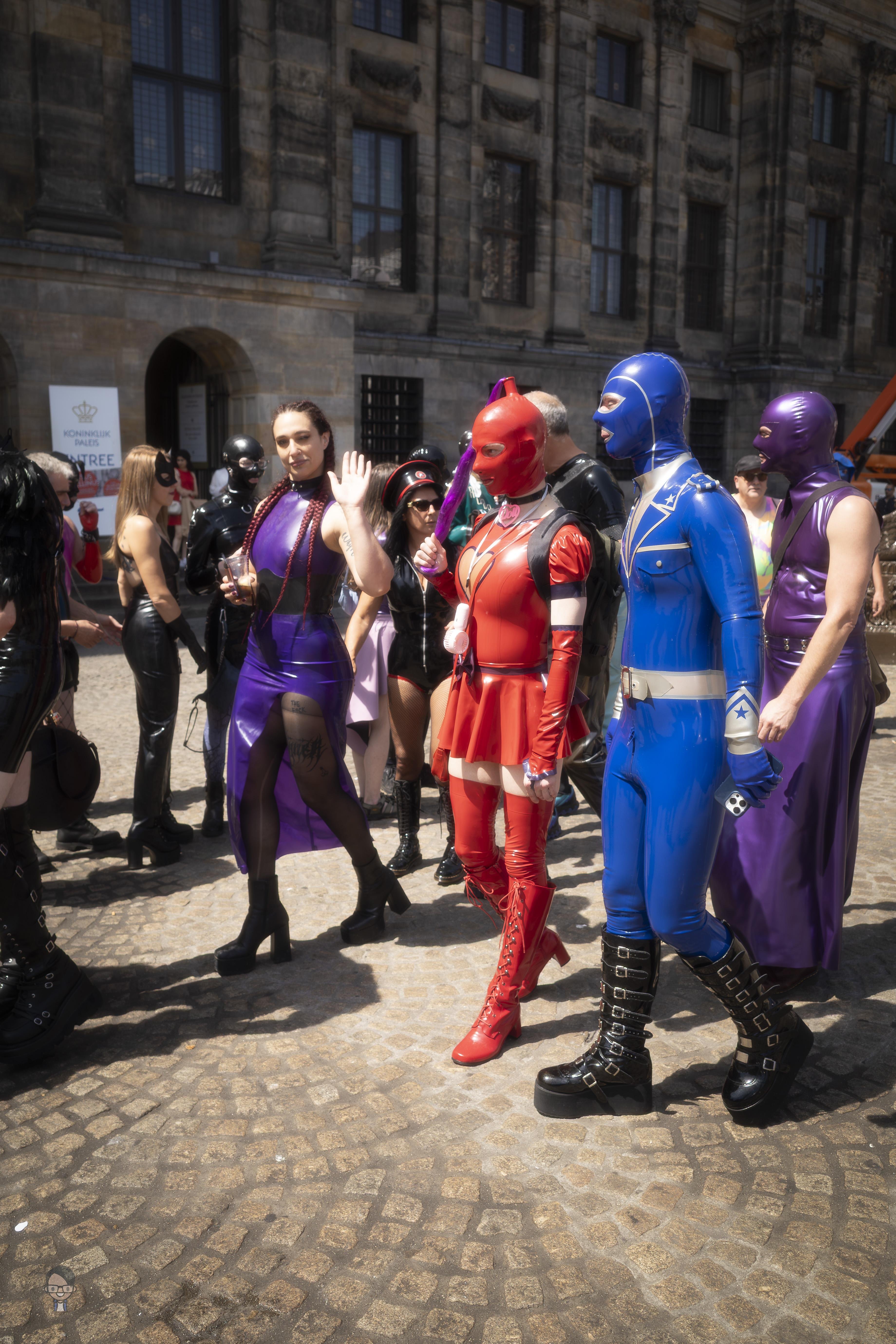 A group of people dressed in colorful costumes featuring latex and leather materials stands on a cobblestone street in front of a historic building. Outfits include a red superhero-style suit with high boots, a blue suit with gloves, and a purple dress with high heels. Some wear masks while others have braided hair. Various accessories, such as a baton and utility belts, are visible. The street is busy with similarly dressed participants, suggesting a themed event or parade.