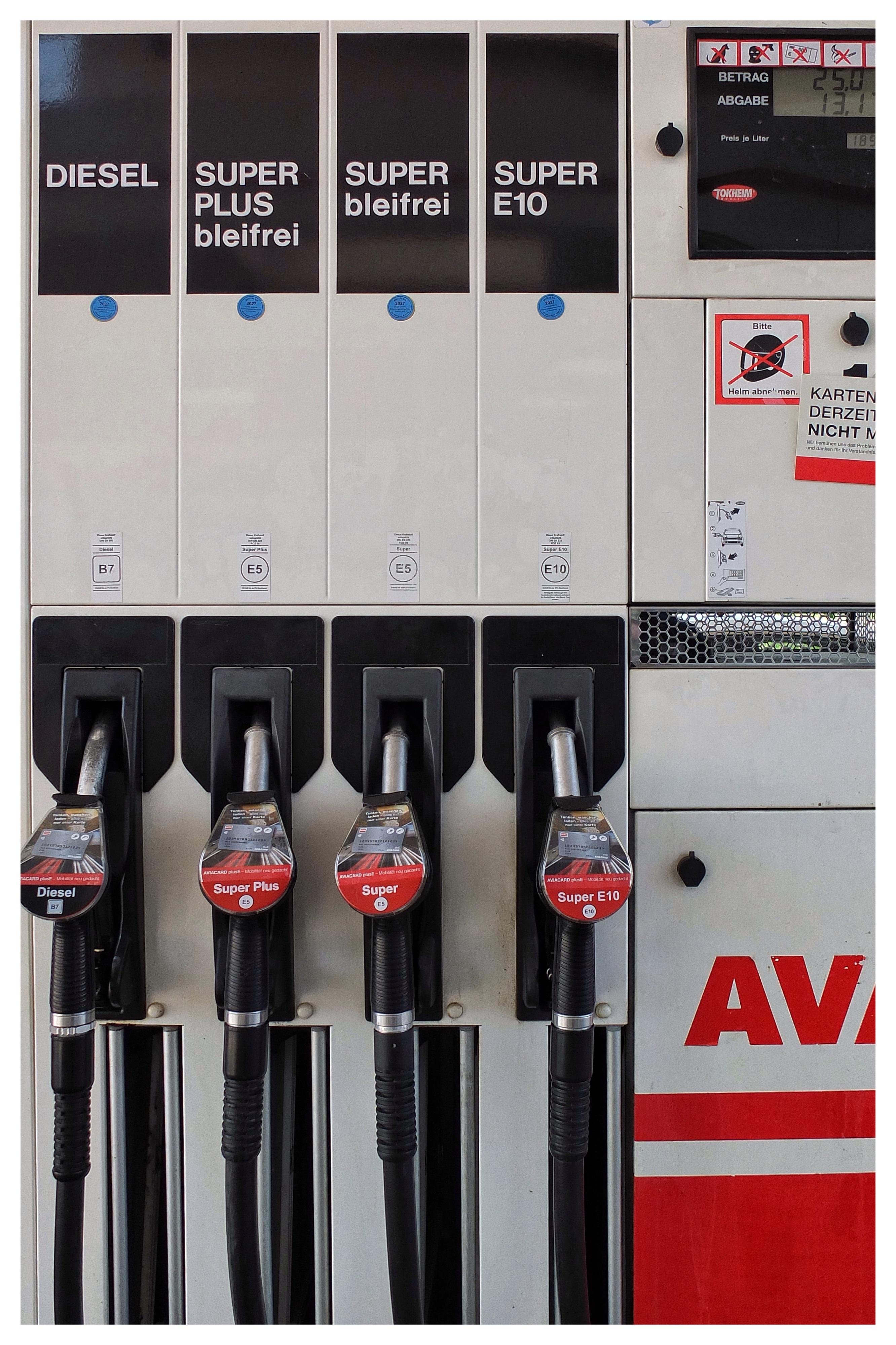 Front view of a gas pump with four fuel options labeled “Diesel,” “Super Plus bleifrei,” “Super bleifrei,” and “Super E10,” each above a corresponding nozzle and hose; a digital payment display and warning stickers are on the right panel.