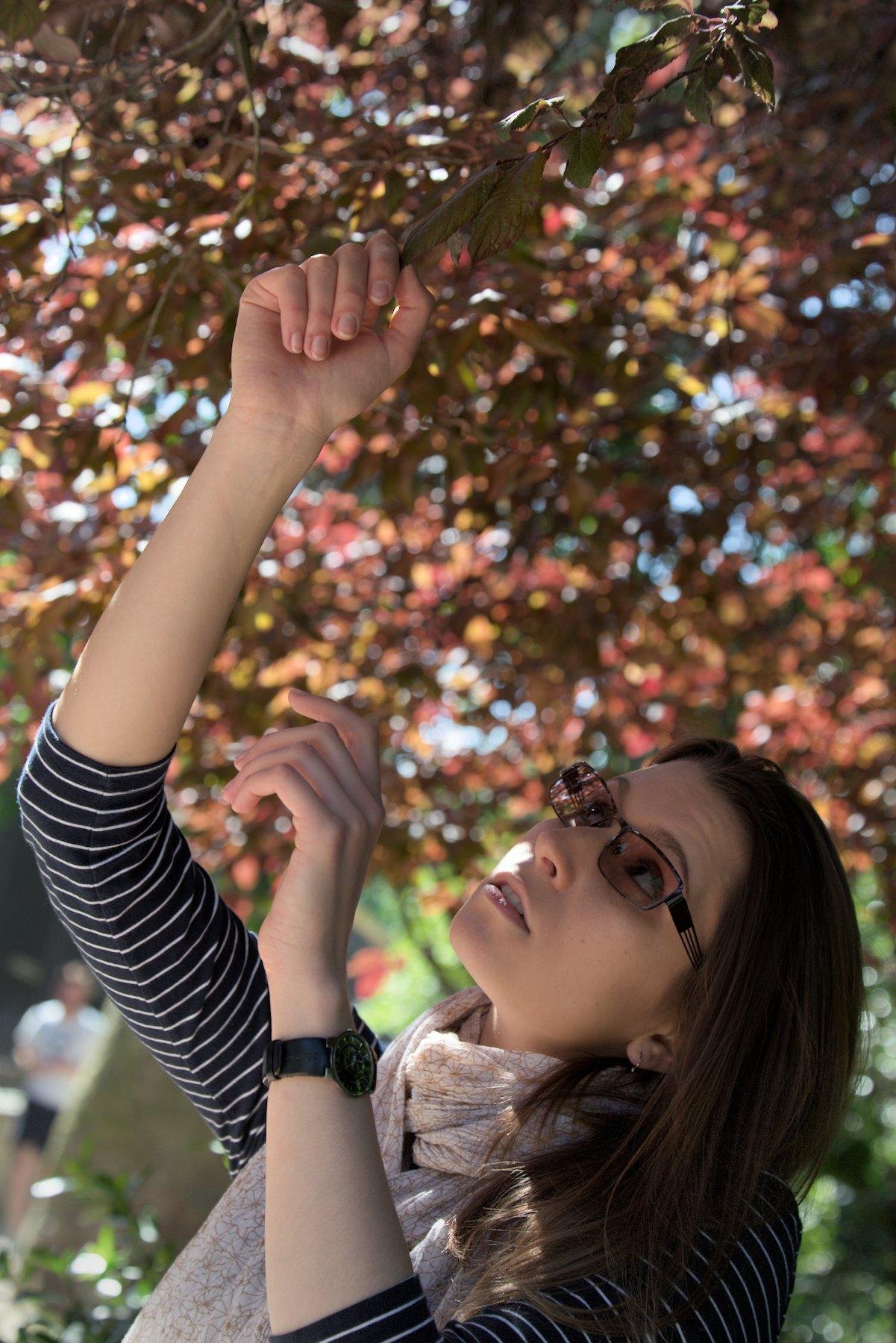 Photo format portrait montrant une jeune femme brune avec des lunettes auto tintantes, une montre noire, un haut bleu marine à rayures blanches horizontales dont les manches vont jusqu’à juste après les coudes. Elle regarde en l’air les feuilles marrons/orangées de l’arbre au dessus d’elle et est en train de saisir une feuille délicatement avec sa main droite, la main gauche étant dans un mouvement pour rejoindre la droite