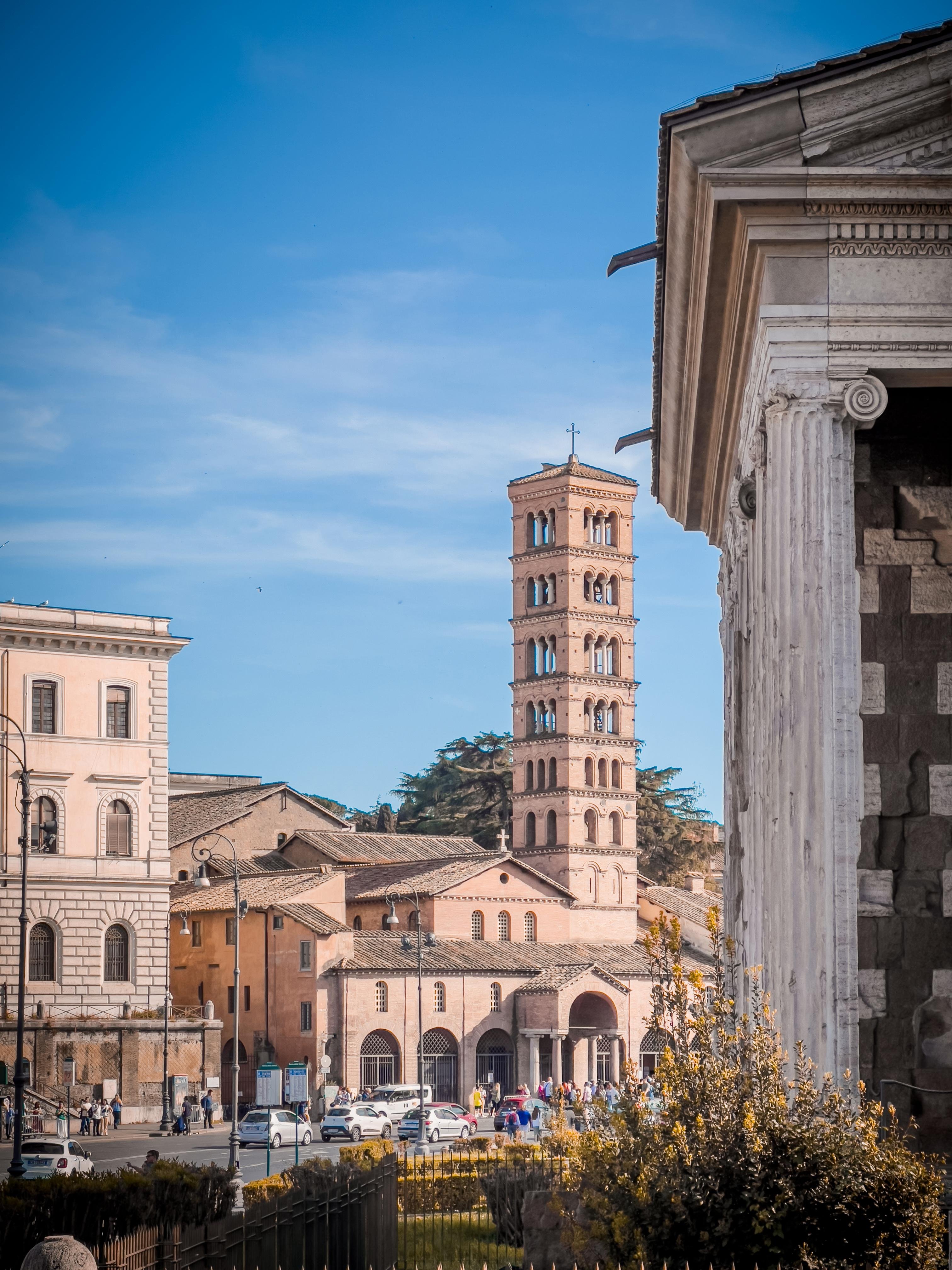Scena urbana con architettura storica. Al centro si erge un alto campanile, probabilmente parte di una chiesa, con più livelli di finestre ad arco. In primo piano a destra, c'è una porzione di un edificio classico con colonne. L'ambiente suggerisce un contesto europeo, possibilmente italiano, dato lo stile architettonico. Molte persone e auto sono visibili, indicando un'area trafficata.