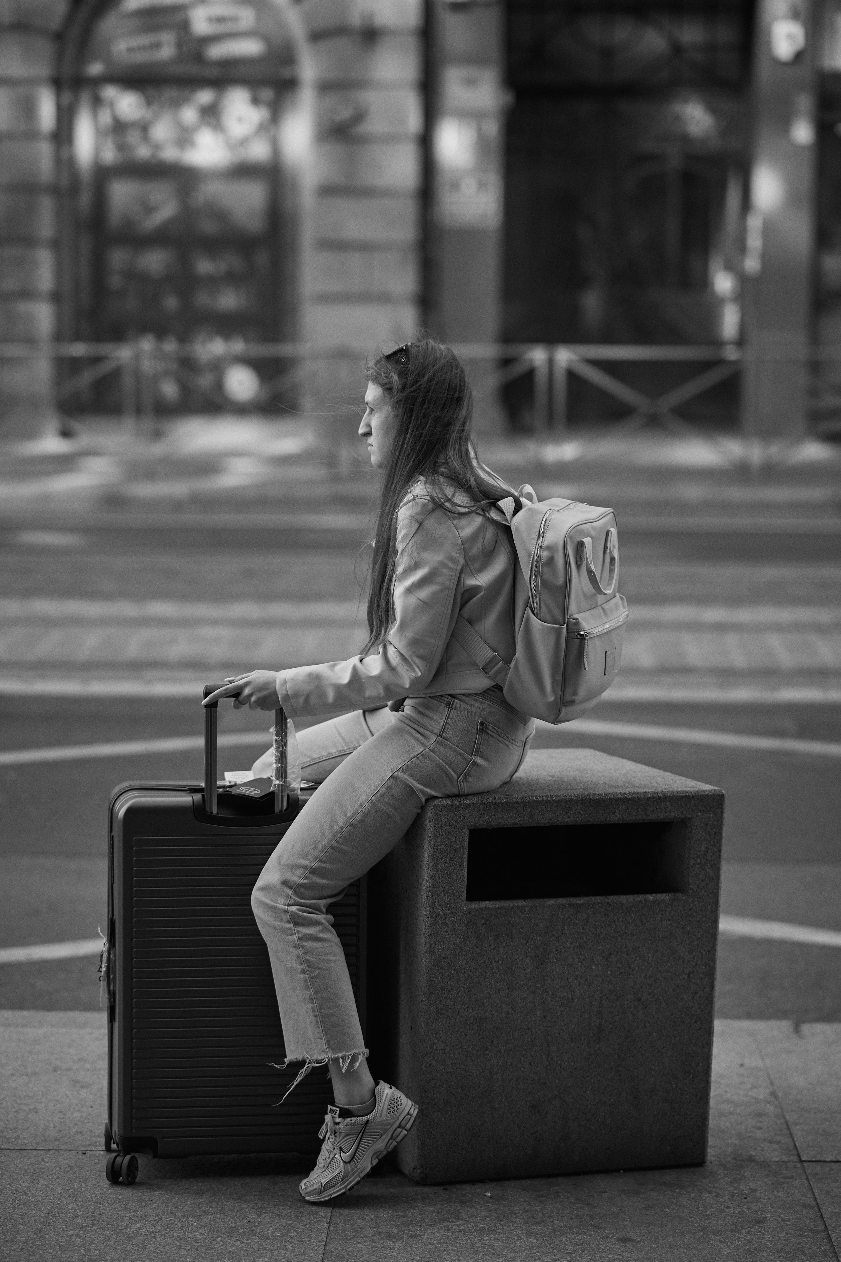 Young woman with long hair sitting on a stone block in a city setting, holding a large wheeled suitcase with one hand. Wears a light-colored jacket, jeans, and sneakers, with a backpack on her back. The background shows a blurred street and building facade. The image is in black and white.