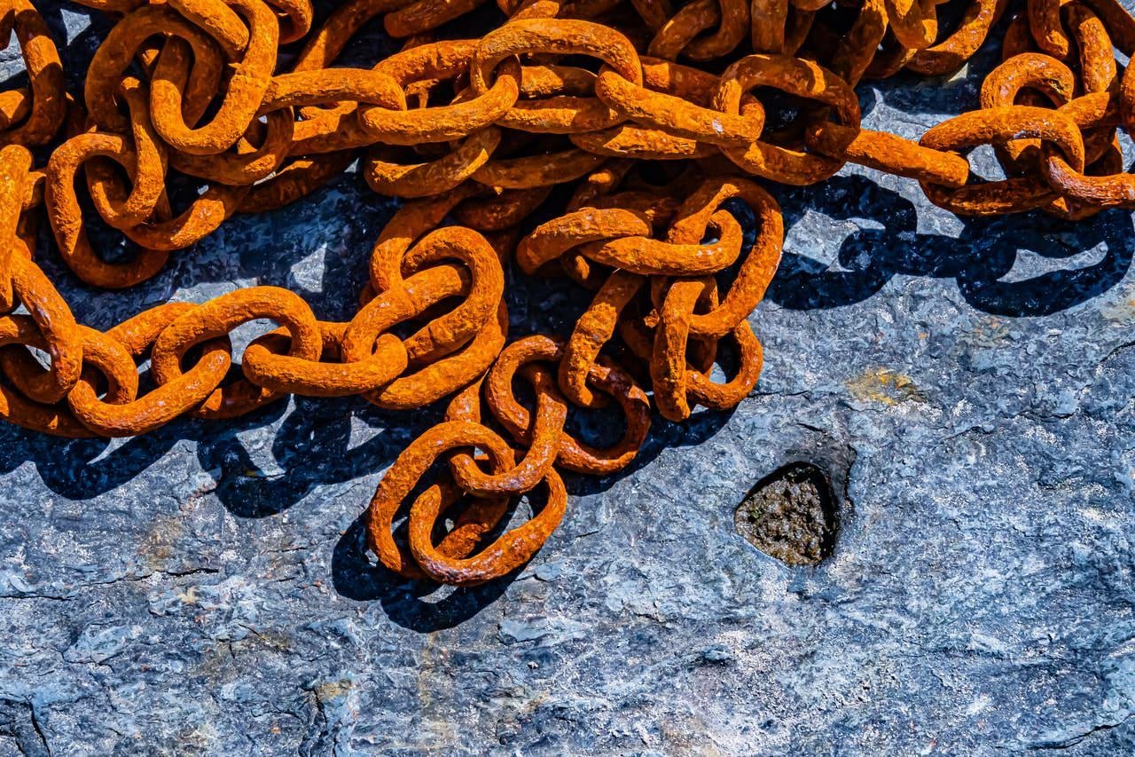 The image shows a pile of rusted metal chains lying on a rough, stone surface. The chains have an orange-brown coloration due to the rust.