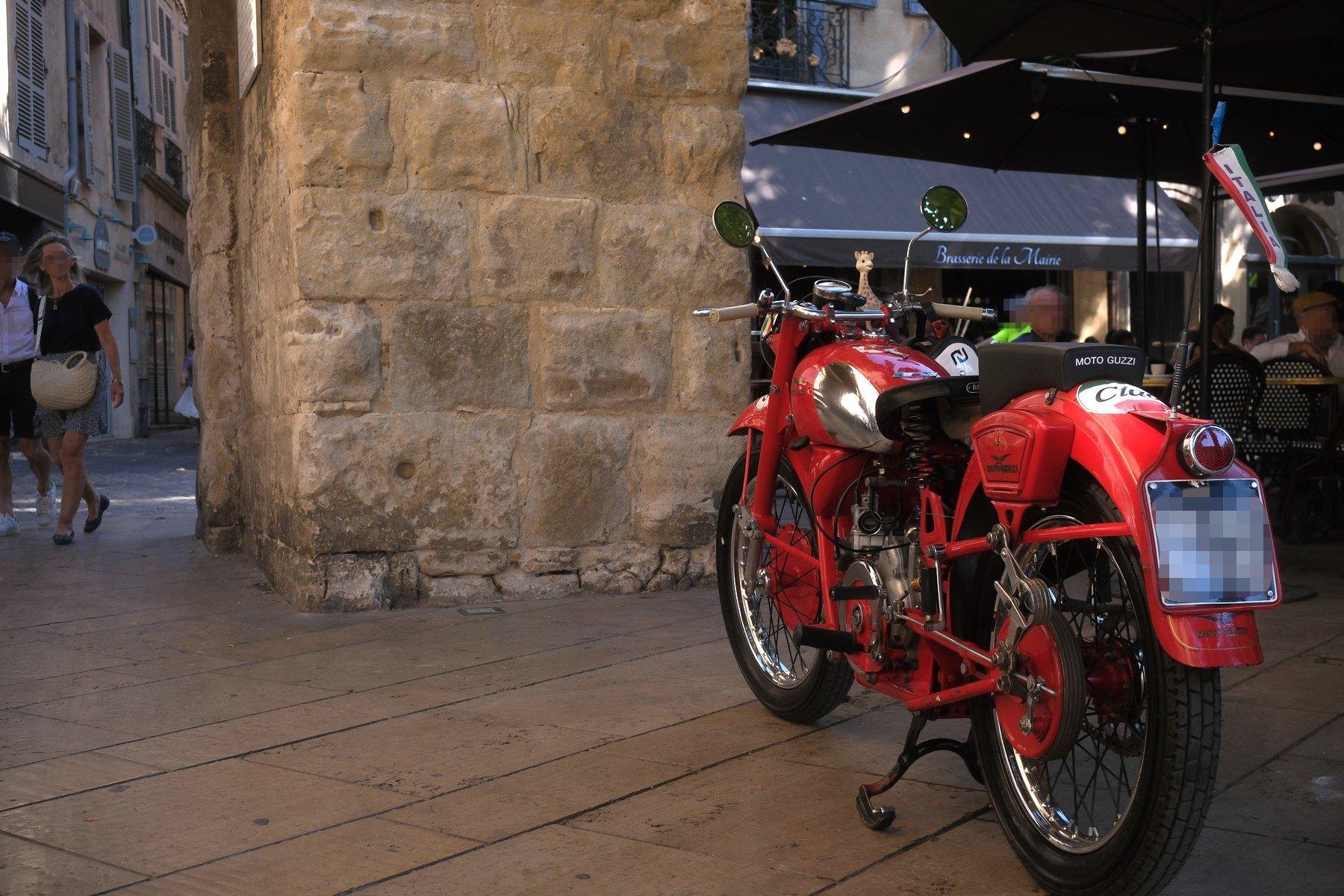 Photo horizontale. Une moto de collection vu de dos, devant un très gros pilier carré et une terrasse de restaurant