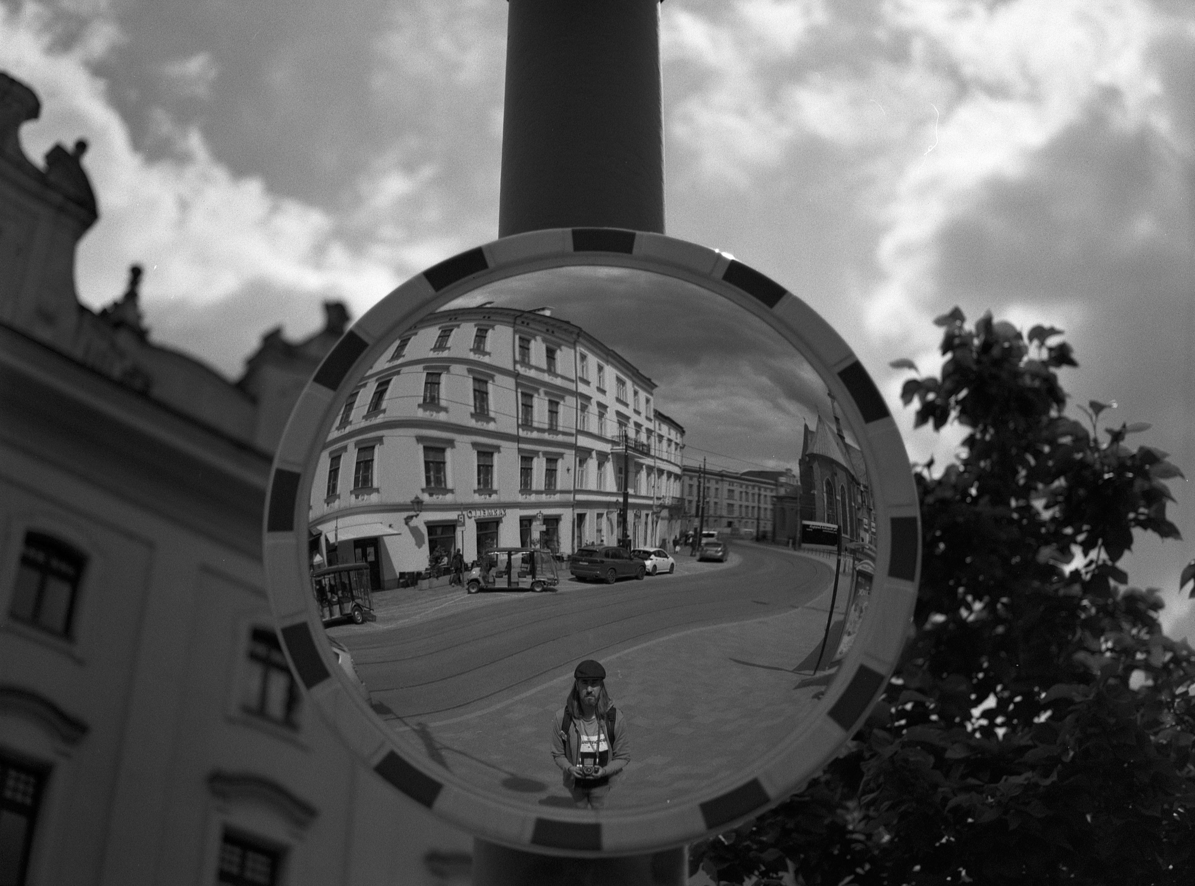 Convex traffic mirror reflecting a street scene with a multi-story building, parked cars, and a person holding a camera standing directly in front. The mirror is mounted on a pole, with a cloudy sky and part of a building and tree visible in the background. The image is in black and white.