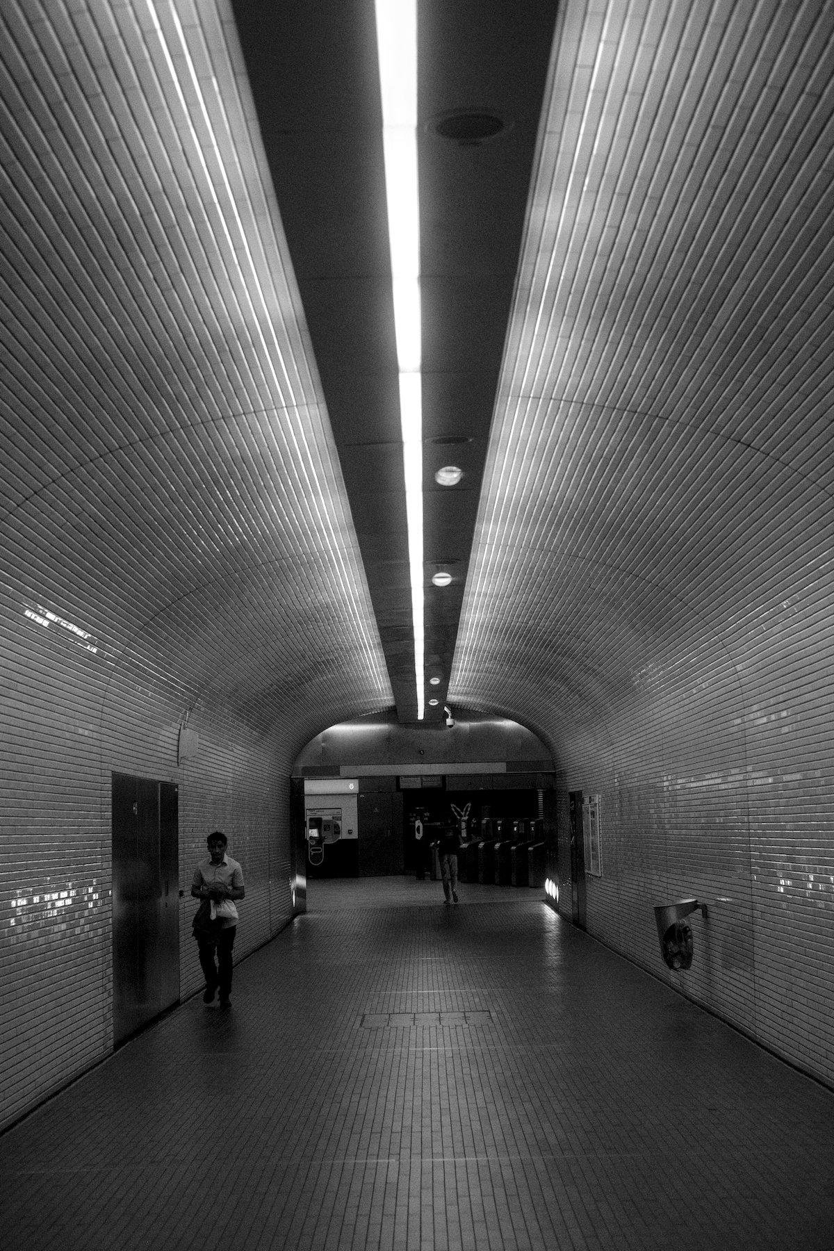 Photo verticale en noir et blanc. Un long tunnel piéton menant aux accès vers le métro. Le plafond a une longue ligne blanche de lumière. Le reste est fait de carrelage caractéristique du métro parisien. Sur la gauche une personne marche vers le premier plan