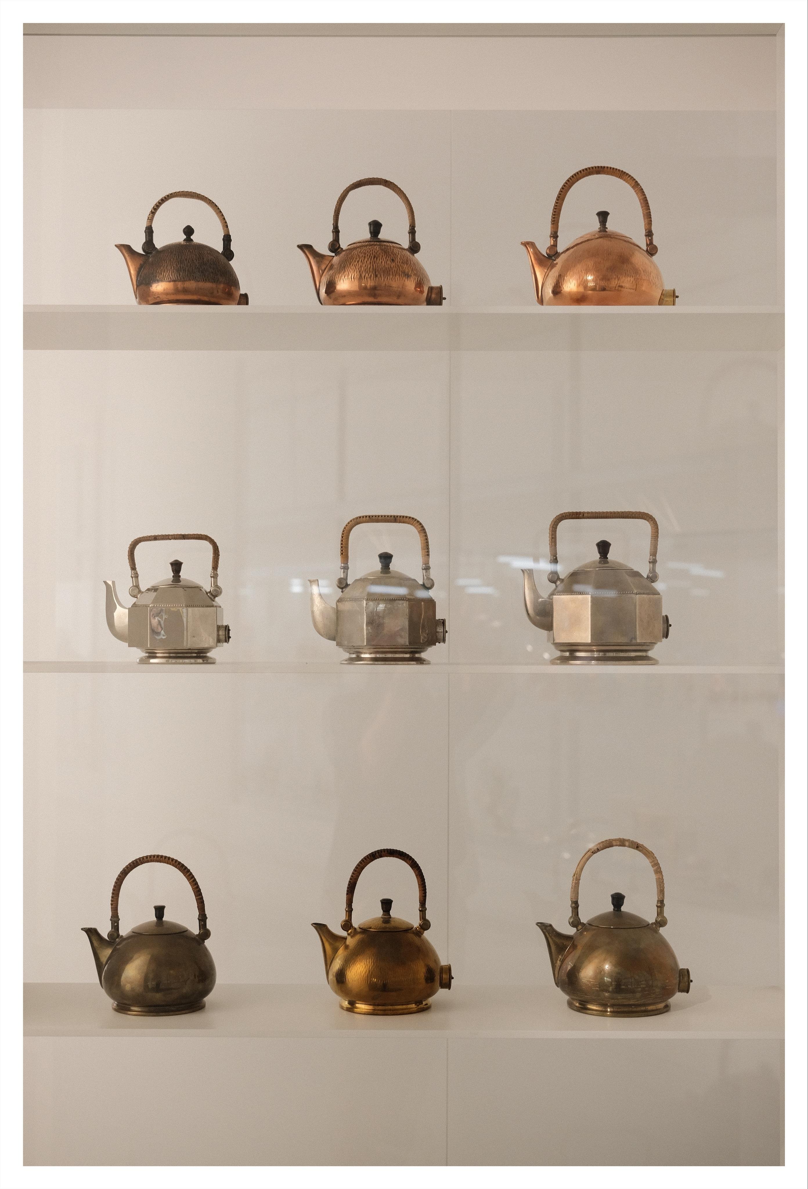 Nine vintage metal kettles displayed in a 3×3 grid on white shelves, with copper kettles on the top row, three faceted silver kettles in the middle row, and darker brass kettles on the bottom row, each with a curved handle and side spout.