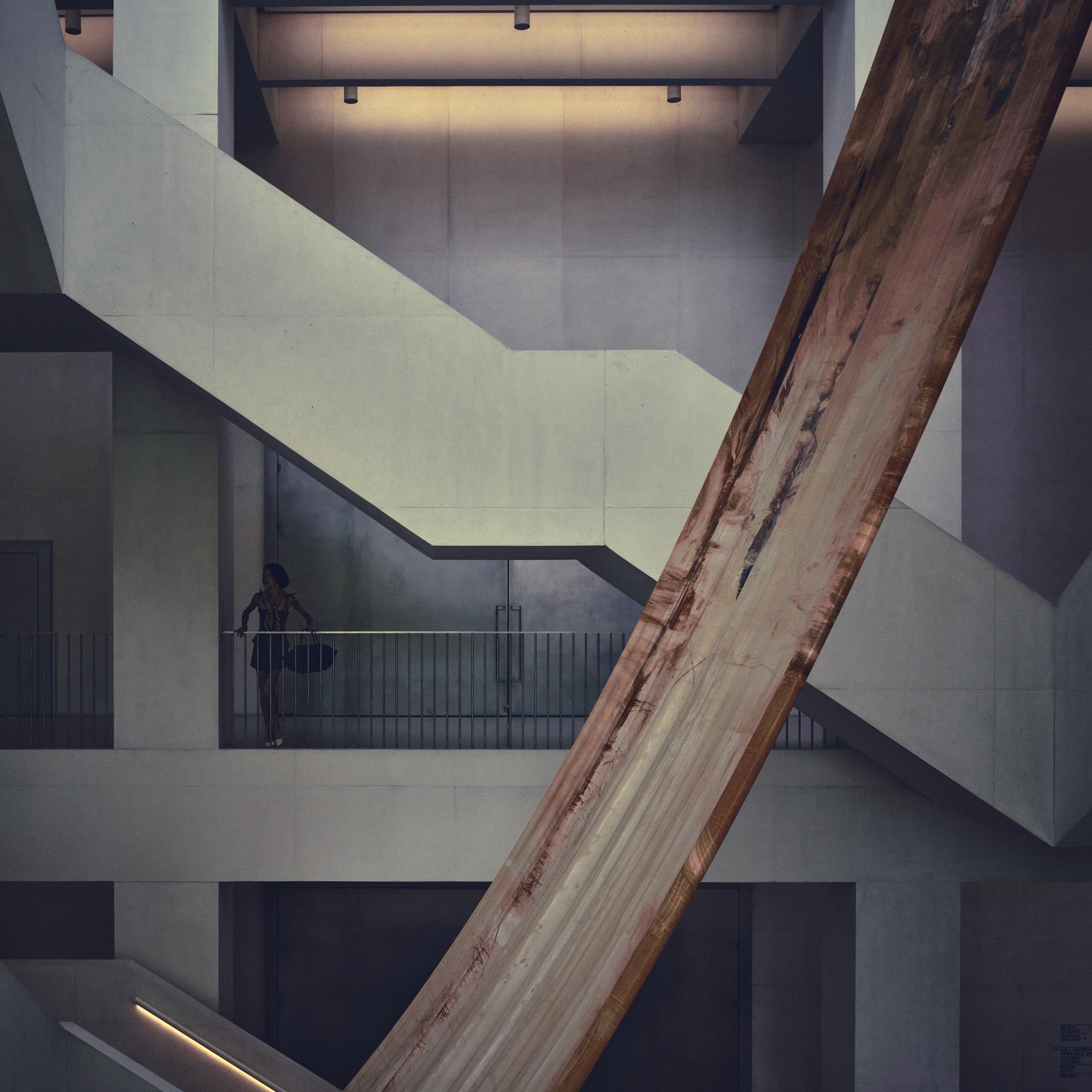 Interior of a modern concrete building with intersecting staircases and railings; a person stands on an upper balcony at left, while a large diagonal wooden beam dominates the foreground.