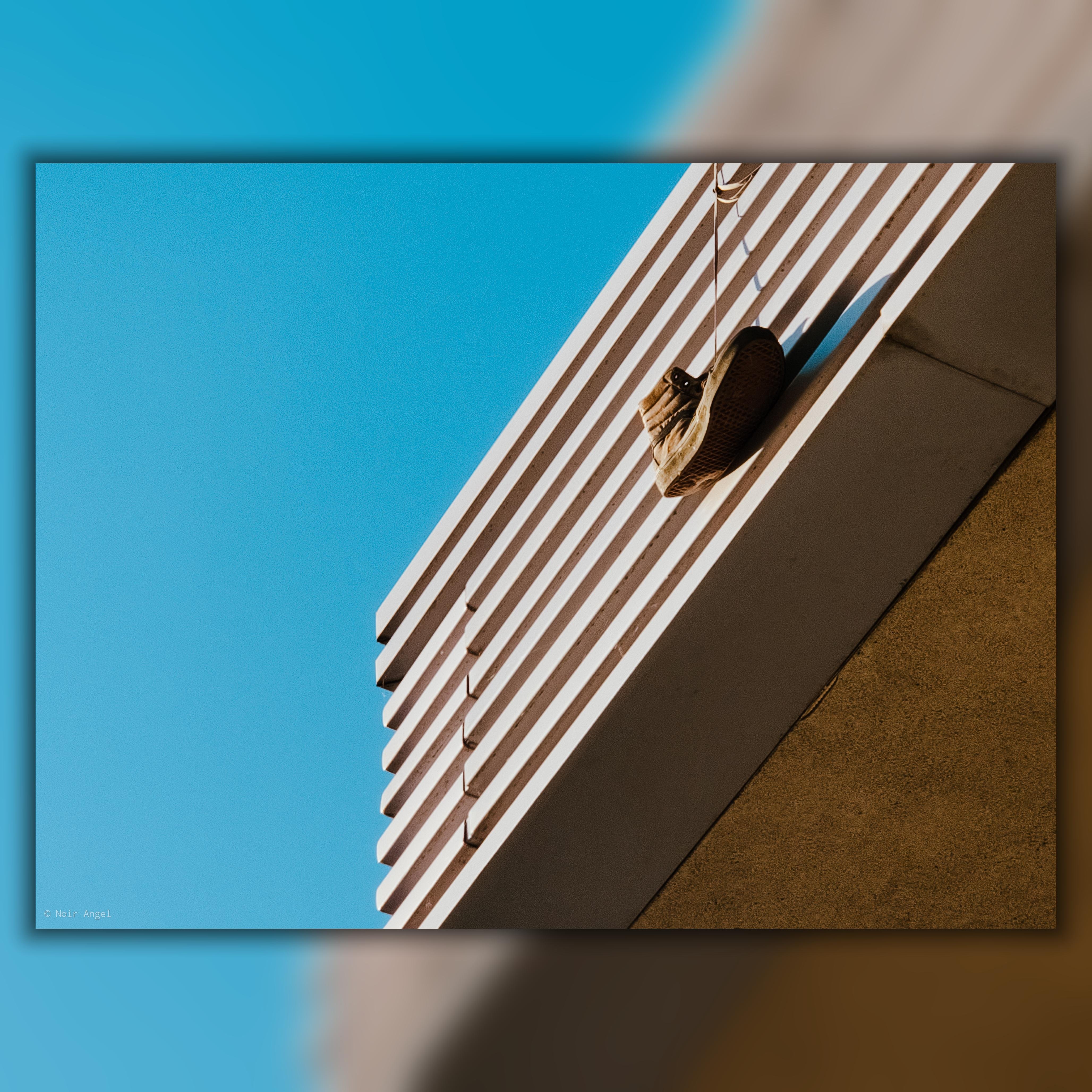 A single brown shoe hanging by its laces from the edge of a building with striped architectural details; set against a clear blue sky. The building has a modern design with horizontal lines.