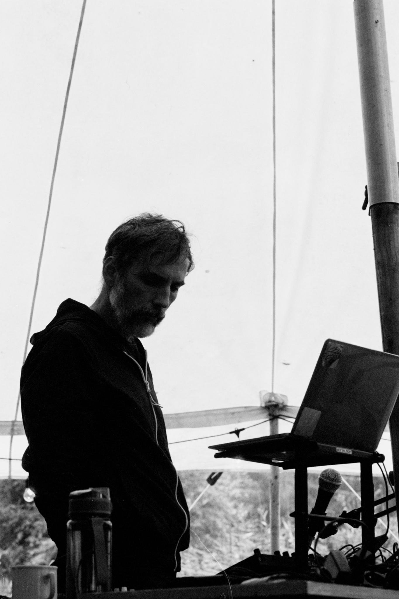Black-and-white photo of a bearded man in a hoodie standing in profile beside a laptop on a stand, with a microphone and audio equipment on a table in the foreground and tent poles and cables in the background.