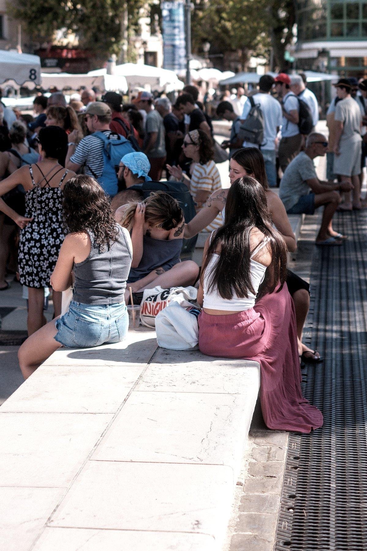 Photo verticale. Sur un muret,  4 jeunes femmes sont assises, l’une d’elle aide son amie se tenant les cheveux, à mettre de la crème solaire dans le dos