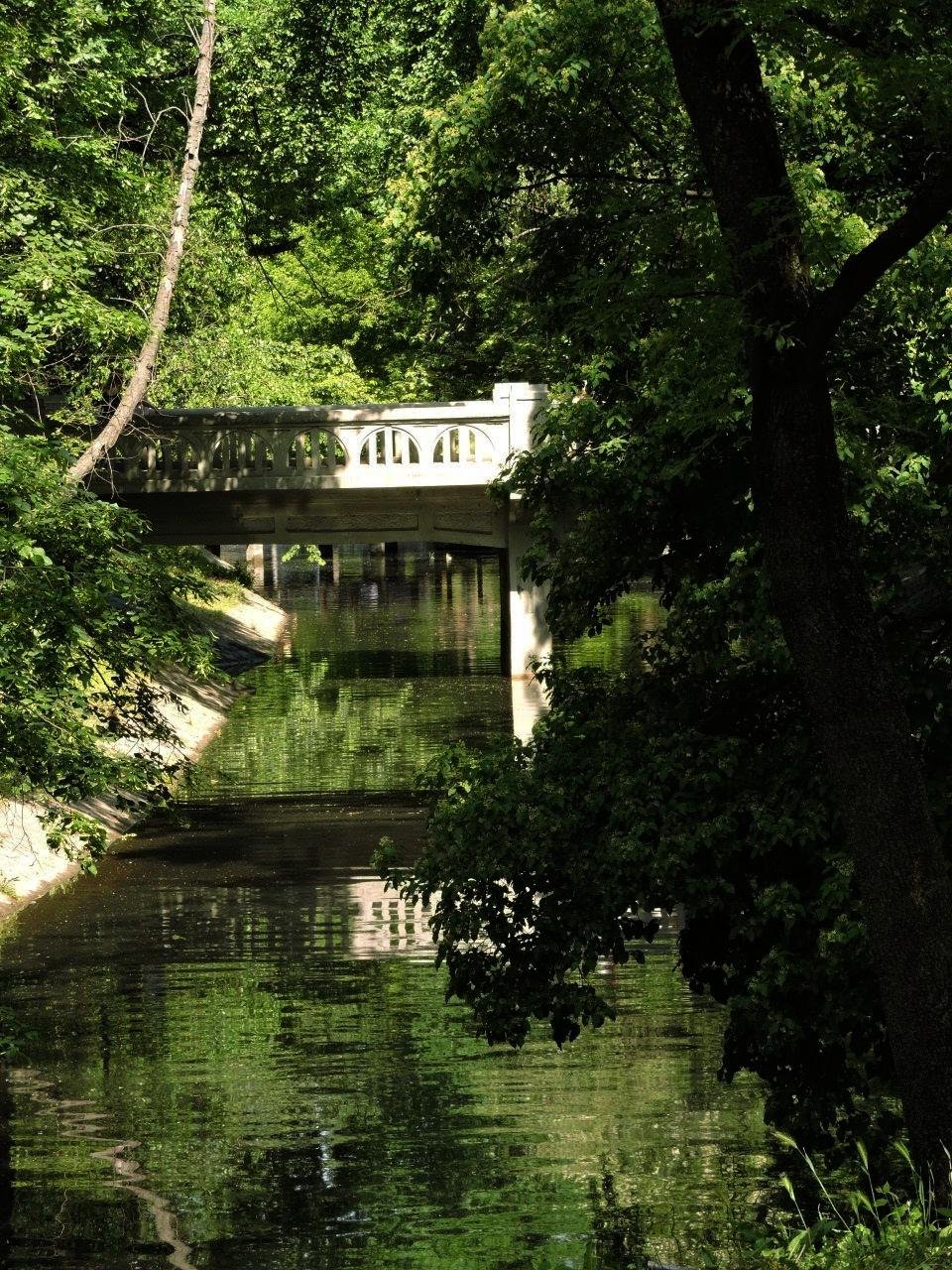 Stone bridge with arched railings crossing over a calm stream surrounded by dense green foliage. Shadows and reflections create patterns on the water's surface. Tall trees frame the scene, with sunlight filtering through the leaves.