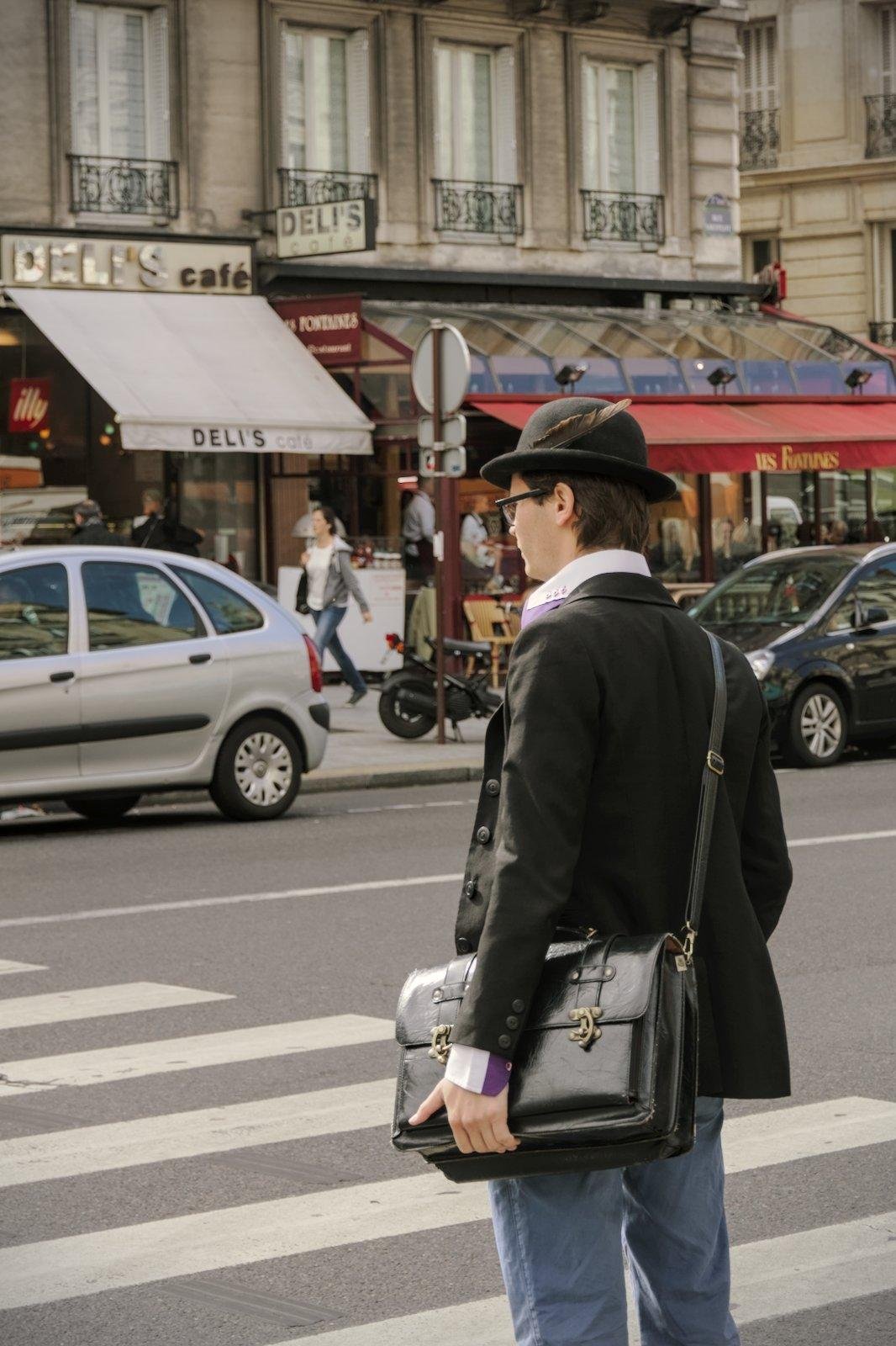 Photo format portrait montrant un homme de dos de 3/4 qui attends à un passage piéton. Il a un chapeau avec une plume fichée dessus, une chemise violette au col blanc et une sacoche en cuir noir