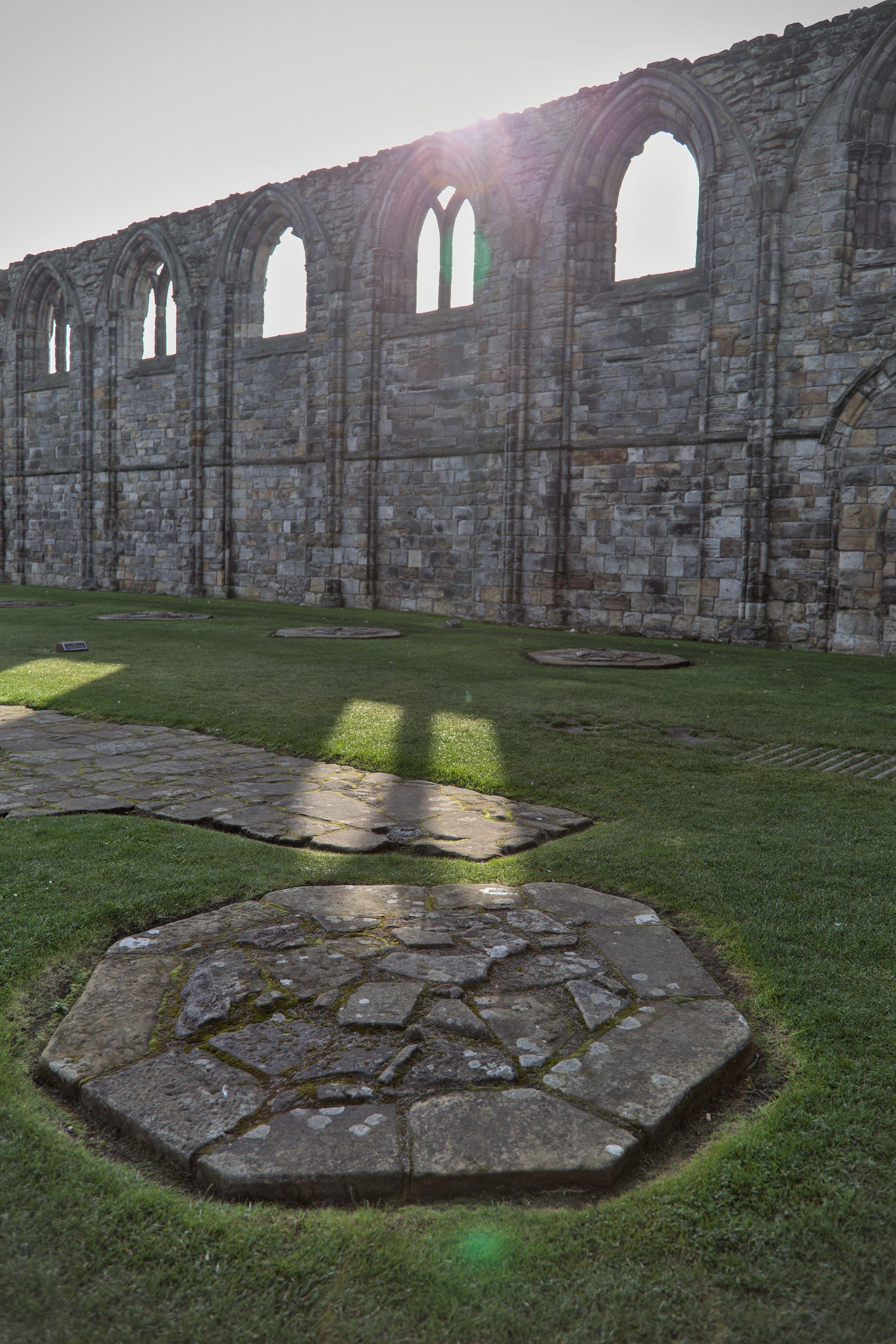 Au premier plan, une colonne en pierre dont il ne reste que la base affleurant le sol. Derrière, les ruines d’un mur en pierre de cathédrale, éclairé par le soleil derrière le mur. Des raies de lumières passent par les «&nbsp;fenêtres&nbsp;» du mur et éclairent la pelouse qui recouvre désormais le sol de l’ancienne cathédrale