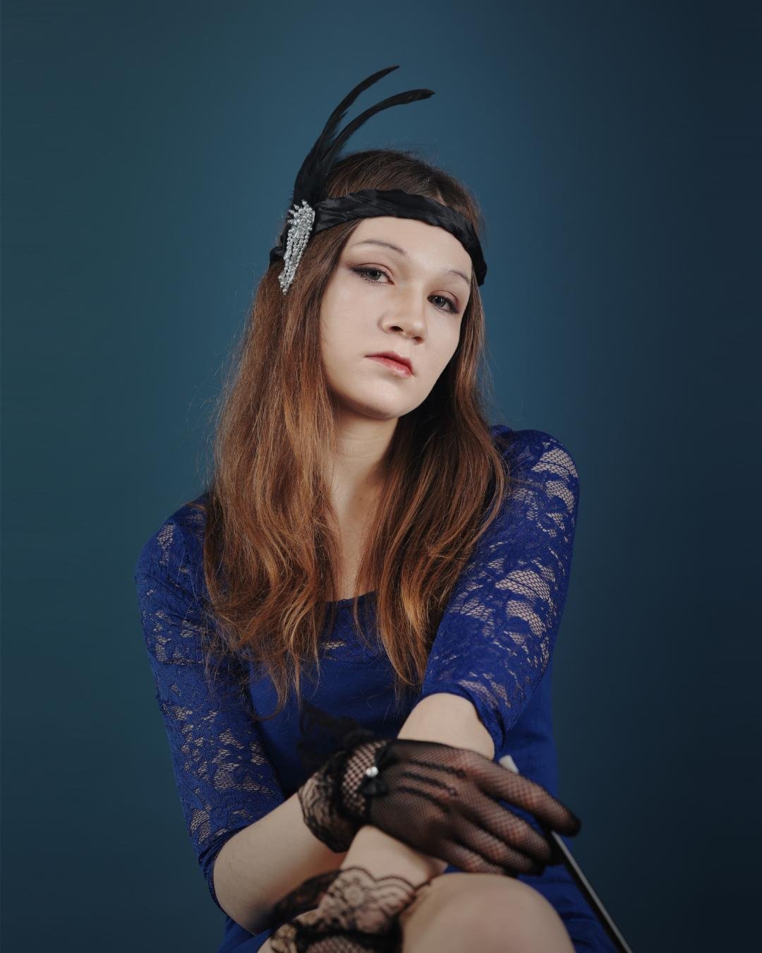 The image shows a person wearing a blue lace dress and a black feathered headband. The individual also has black lace gloves and is posing against a dark background. The attire has a vintage or classic style, reminiscent of the Roaring Twenties.
