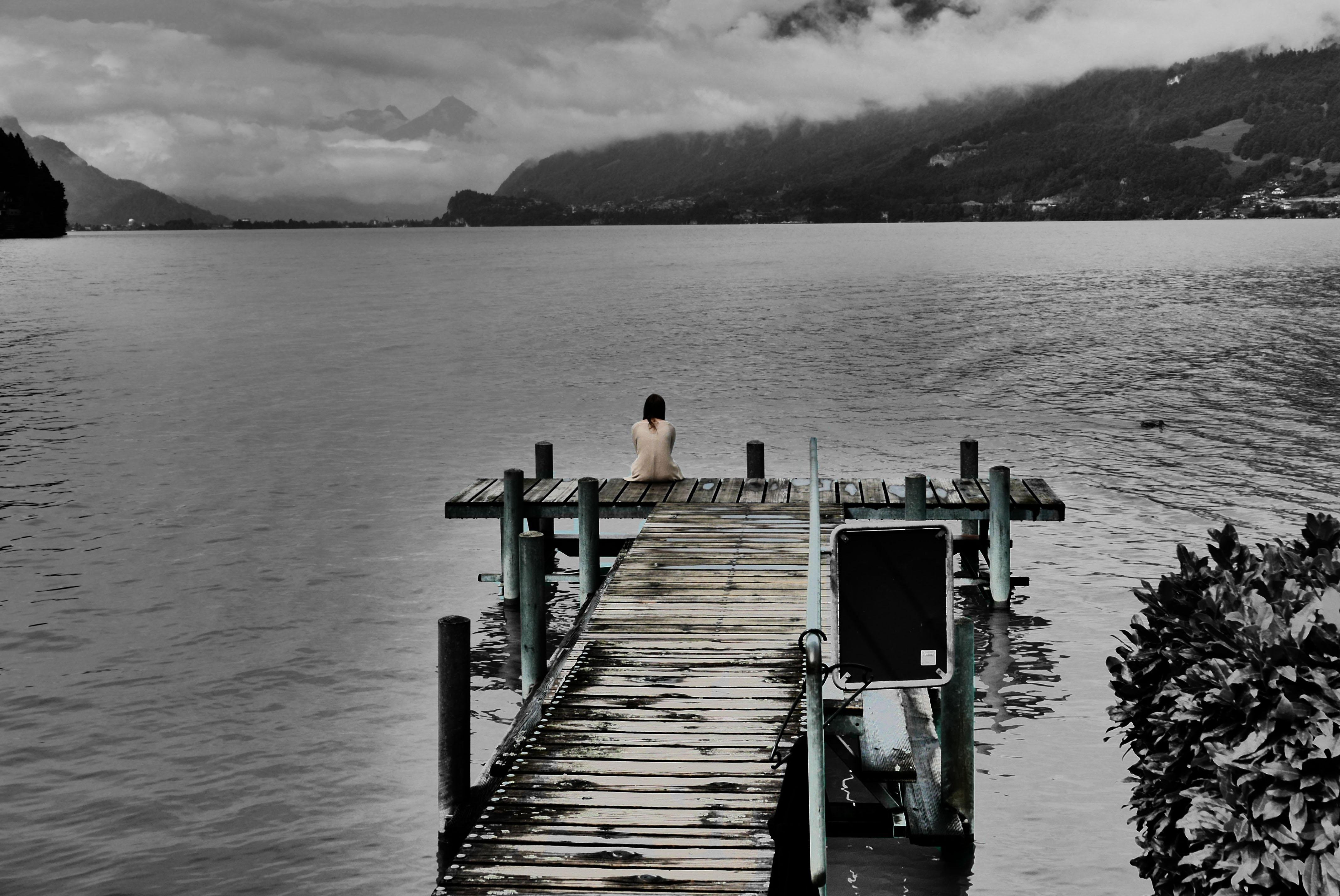 The image features a person sitting alone at the end of a wooden pier, looking out over a large lake. The landscape is serene, with mountains in the distance and clouds above. The image has a subtle color filter, making it largely monochromatic with a muted color tone. The overall atmosphere is calm and reflective.