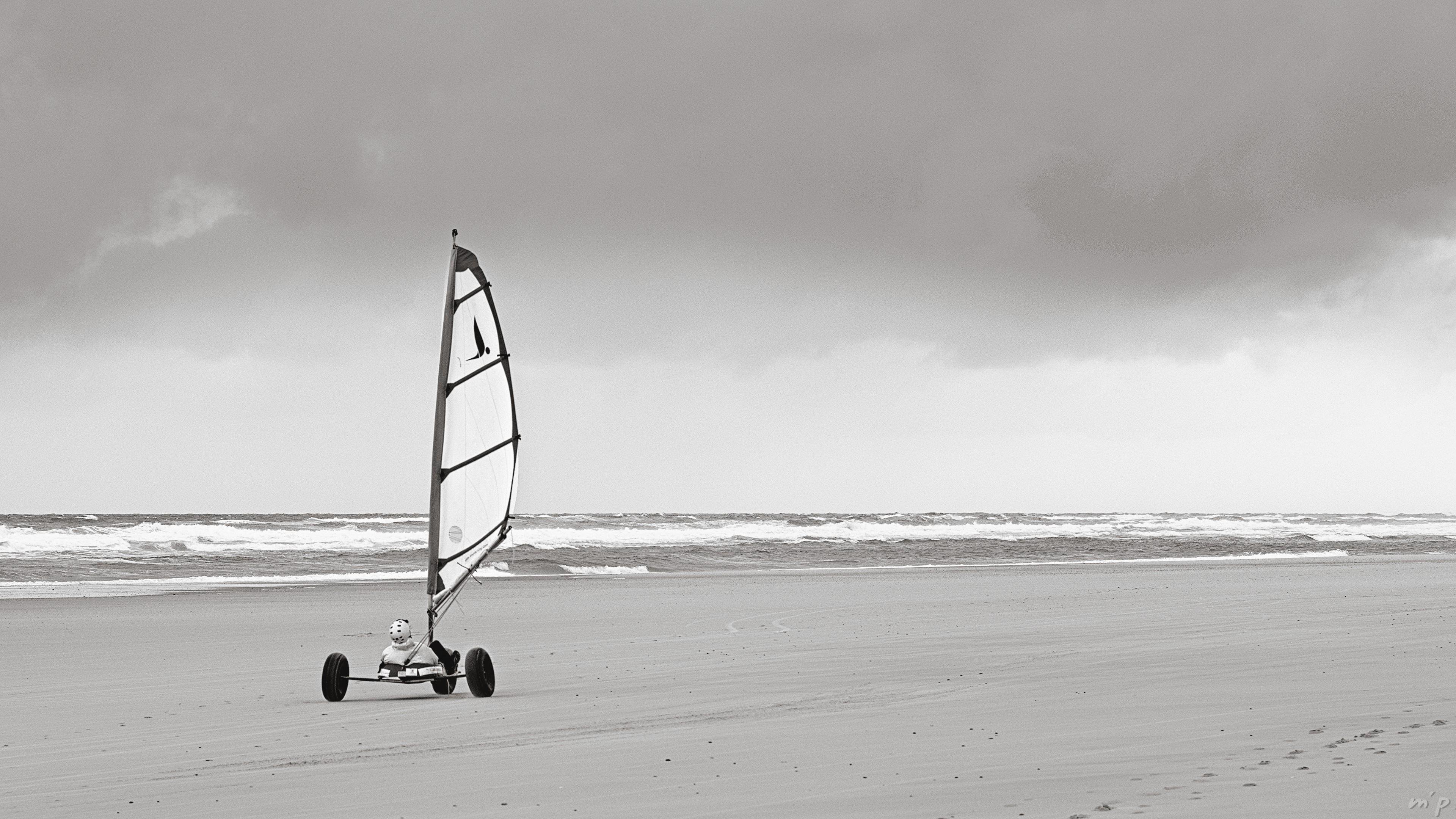 🇬🇧 Horizontal black and white photo of a sand yacht rolling over an ocean beach at low tide under cloudy skies. A stylized silhouette of a bird is placed on the sail of the machine.
///
🇫🇷 Photo noir et blanc horizontale d'un char à voile roulant sur une plage de l'océan à marée basse sous un ciel nuageux. Une silhouette stylisée d'oiseau est plaquée sur la voile de l'engin.