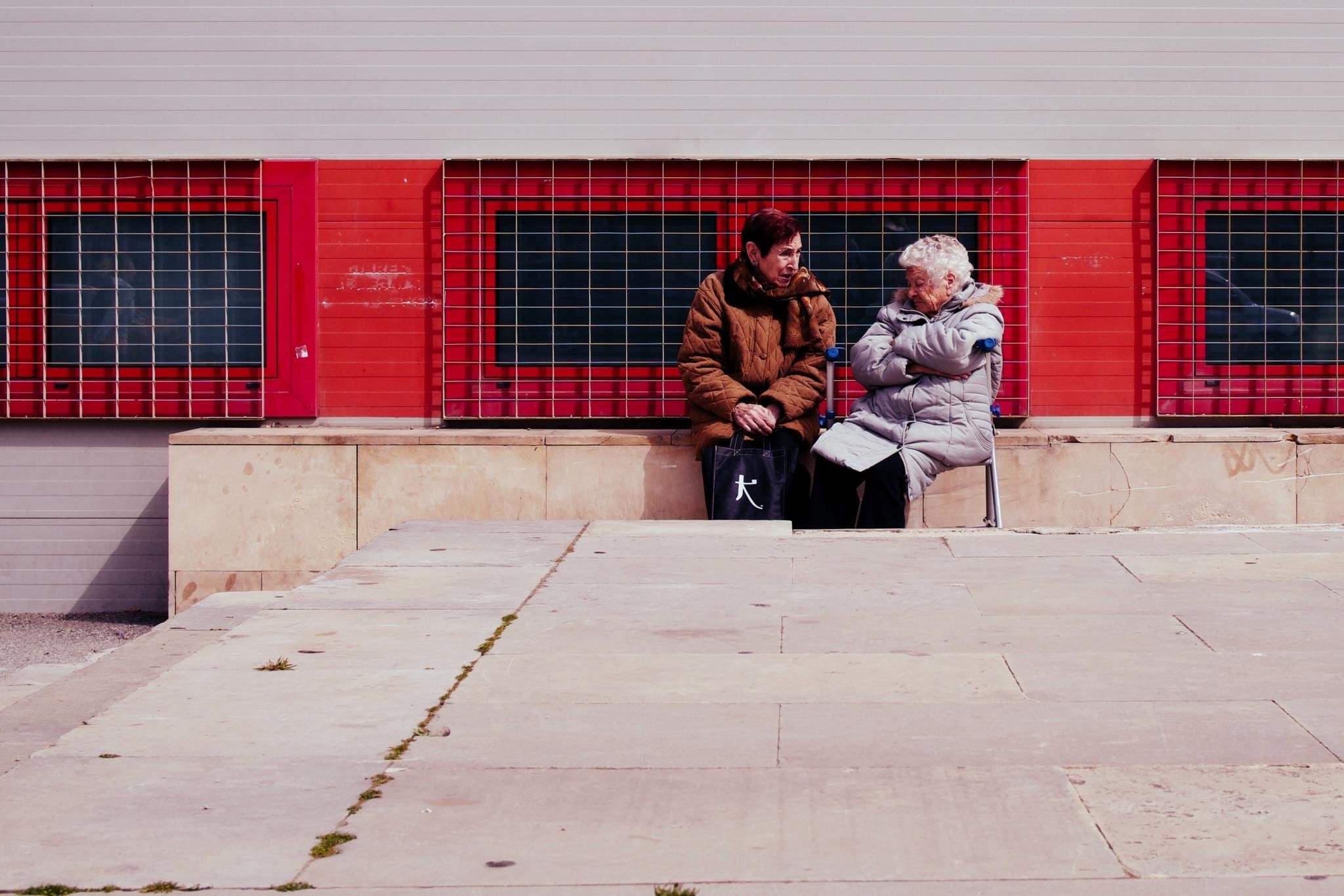 Zwei ältere Menschen sitzen nebeneinander auf Stühlen vor einem roten Gebäude mit vergitterten Fenstern und unterhalten sich; einer trägt einen braunen Mantel und hält eine schwarze Tasche, der andere trägt einen hellgrauen Puffermantel mit verschränkten Armen, mit einem breiten leeren Steinbürgersteig im Vordergrund.