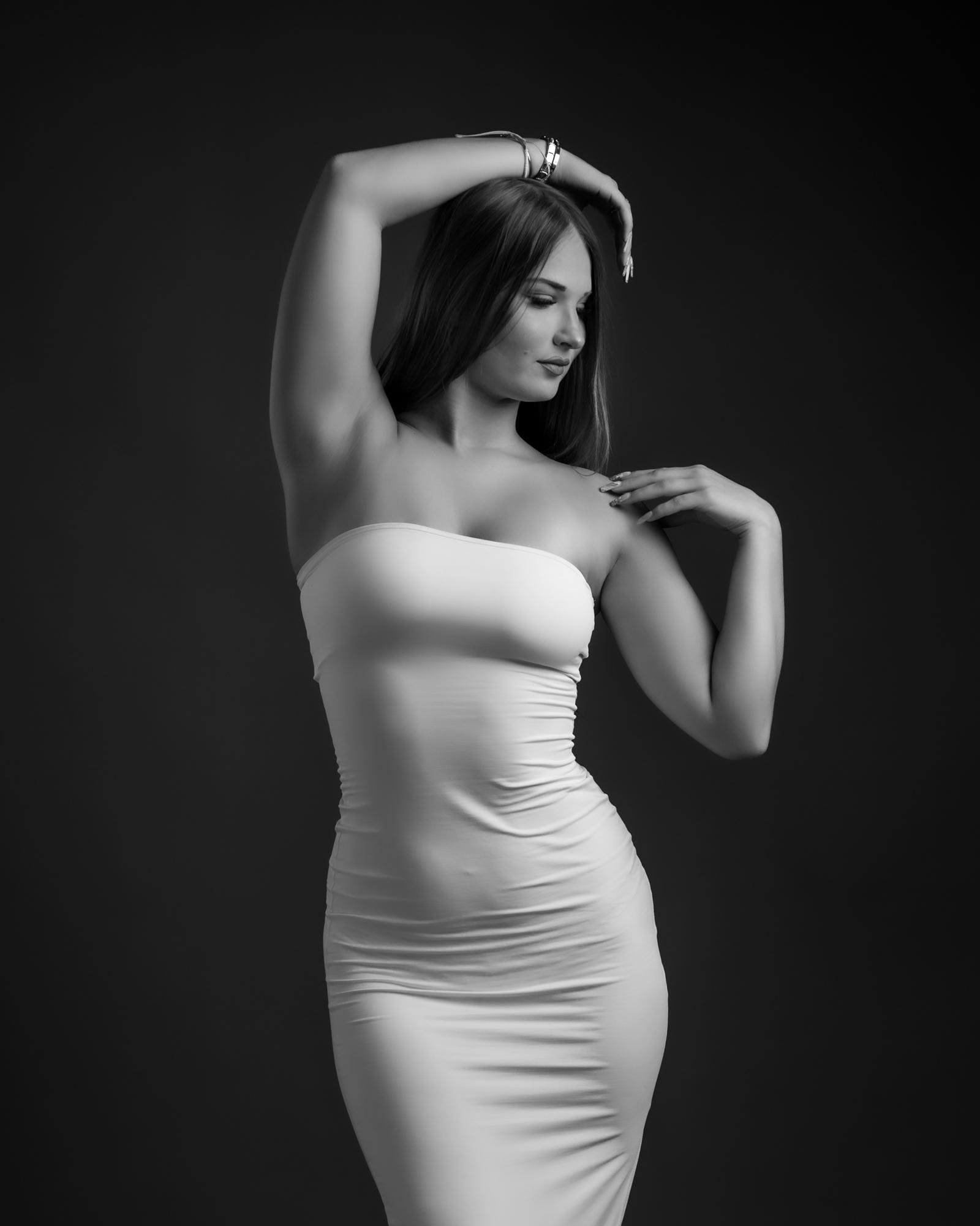 The image shows a black and white photograph of a woman posing elegantly. She is wearing a form-fitting strapless dress and has one arm raised above her head, with the other positioned near her chest. The background is plain and dark, highlighting the subject.