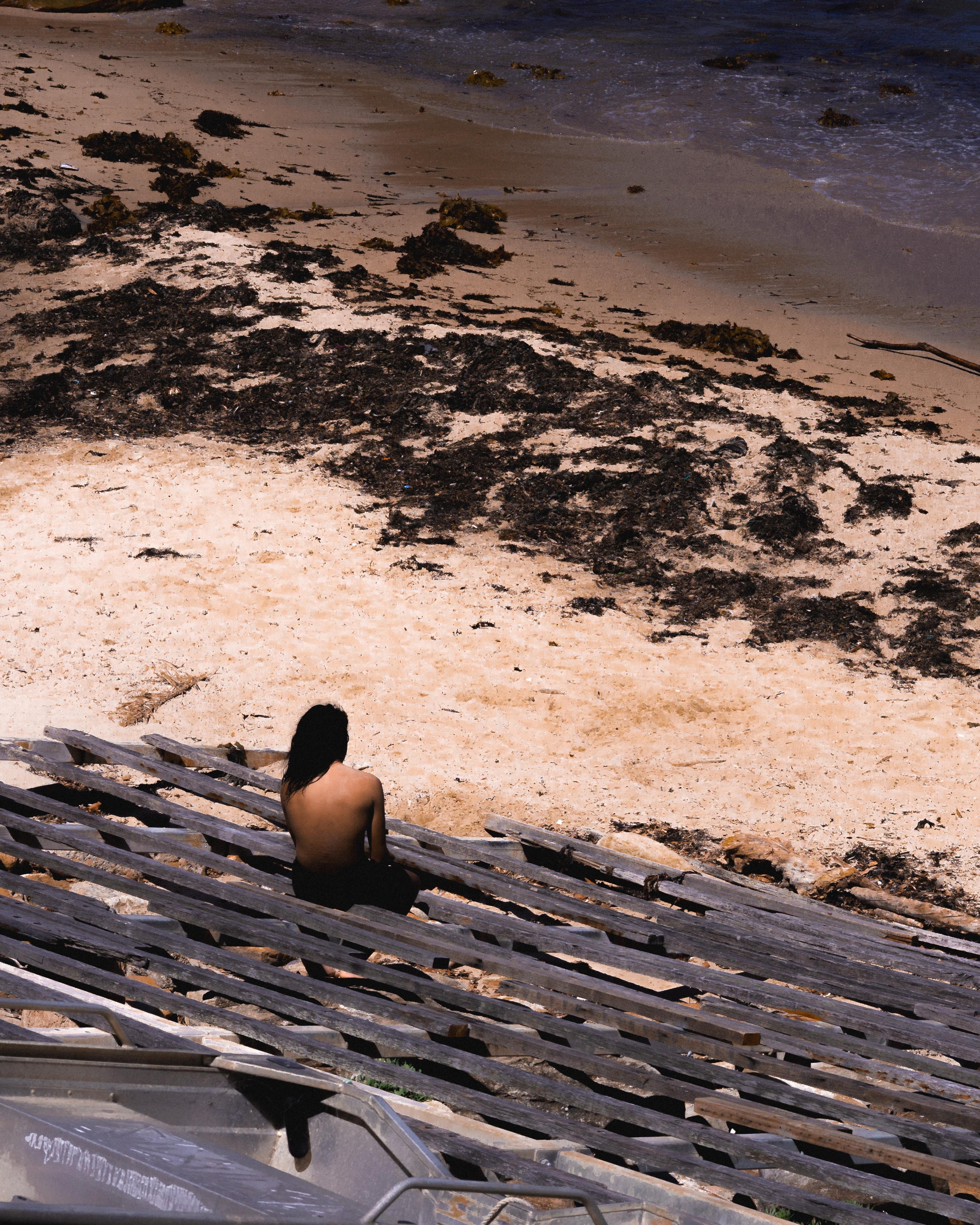 Person with long hair sitting on wooden steps, overlooking a sandy beach with dark patches of seaweed, and gentle waves approaching the shore.