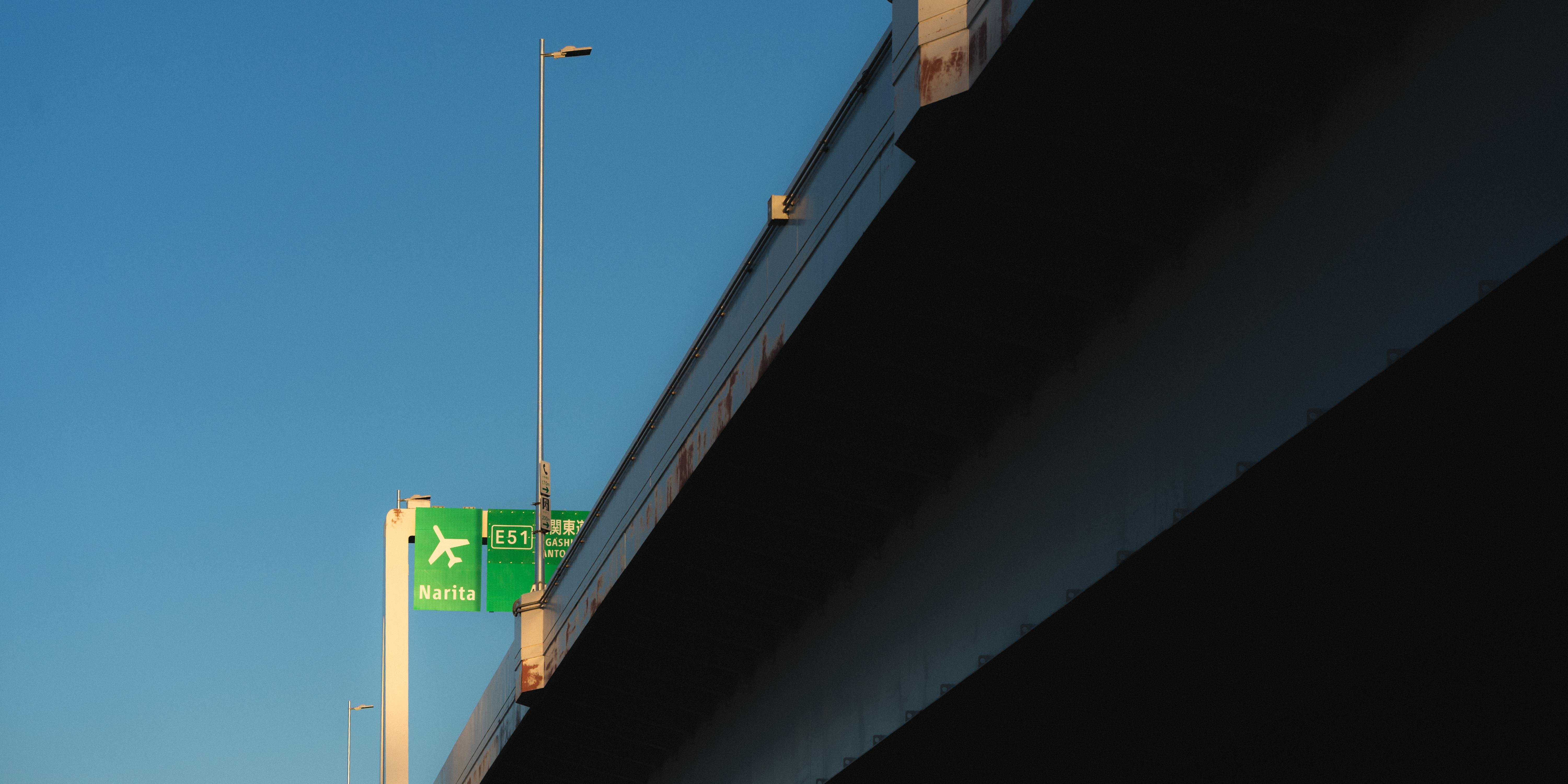 A highway overpass with a clear blue sky in the background. A green road sign attached to the overpass displays an airplane symbol with the text "Narita" and "E51". Two tall lampposts extend from the overpass, casting minimal shadow. The image captures a section of the overpass structure in shadow on the right.