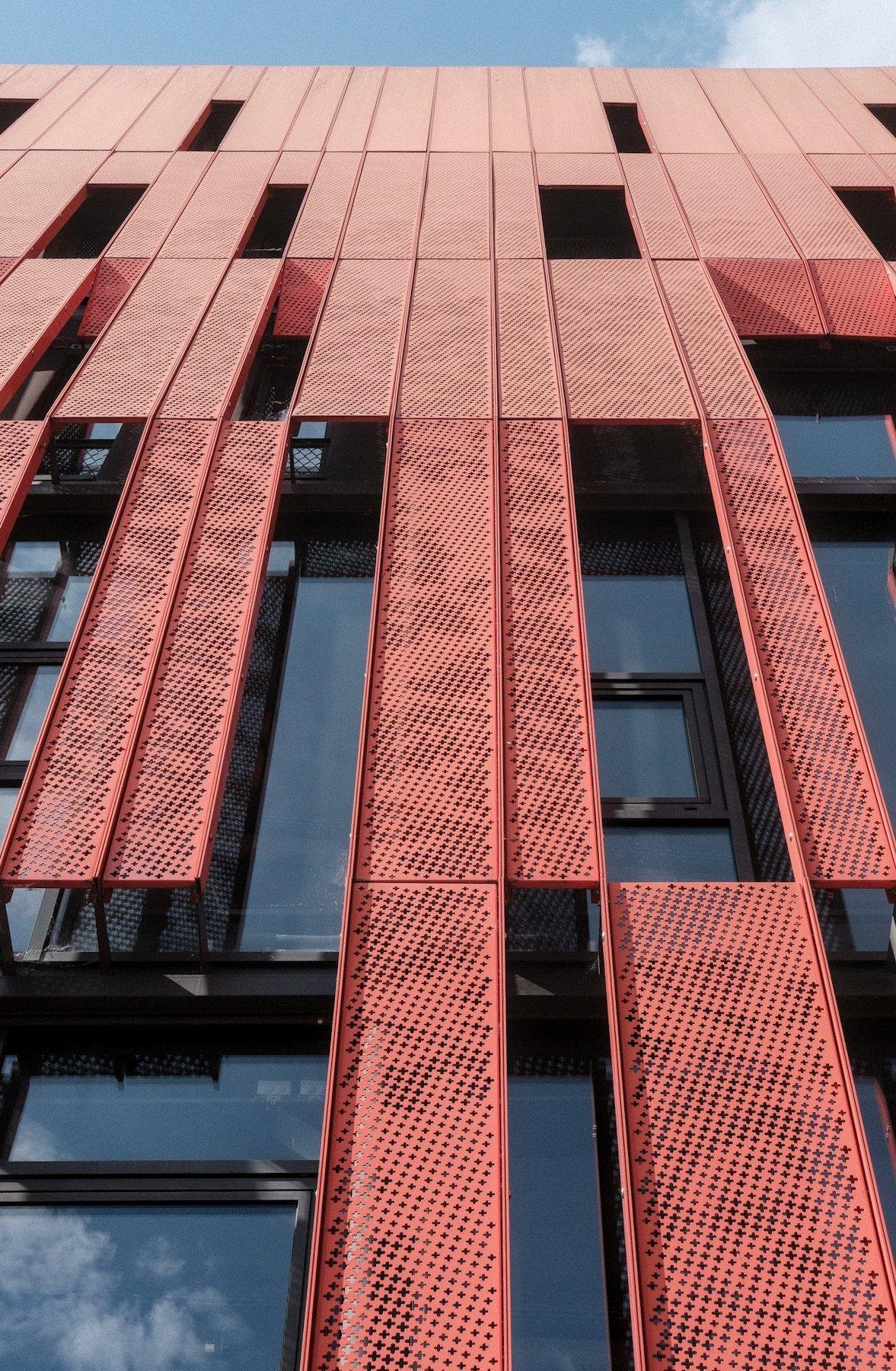 Photo verticale. Contre-plongée sur un bâtiment moderne fait de verre et couvert de bandes de métal trouées peintes en rouge, placée de manière un peu aléatoire, formant comme des rampes menant au ciel bleu