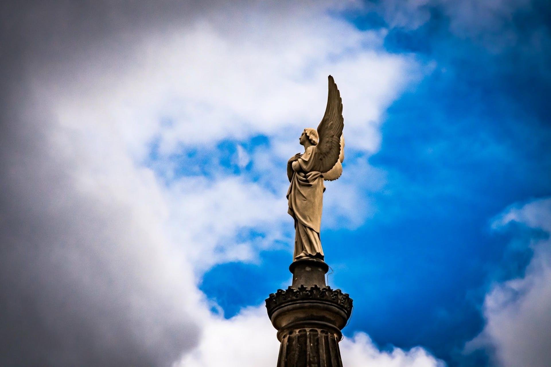 Stone angel statue with one wing raised, standing atop an ornate column, framed against a blue sky with large white and gray clouds.