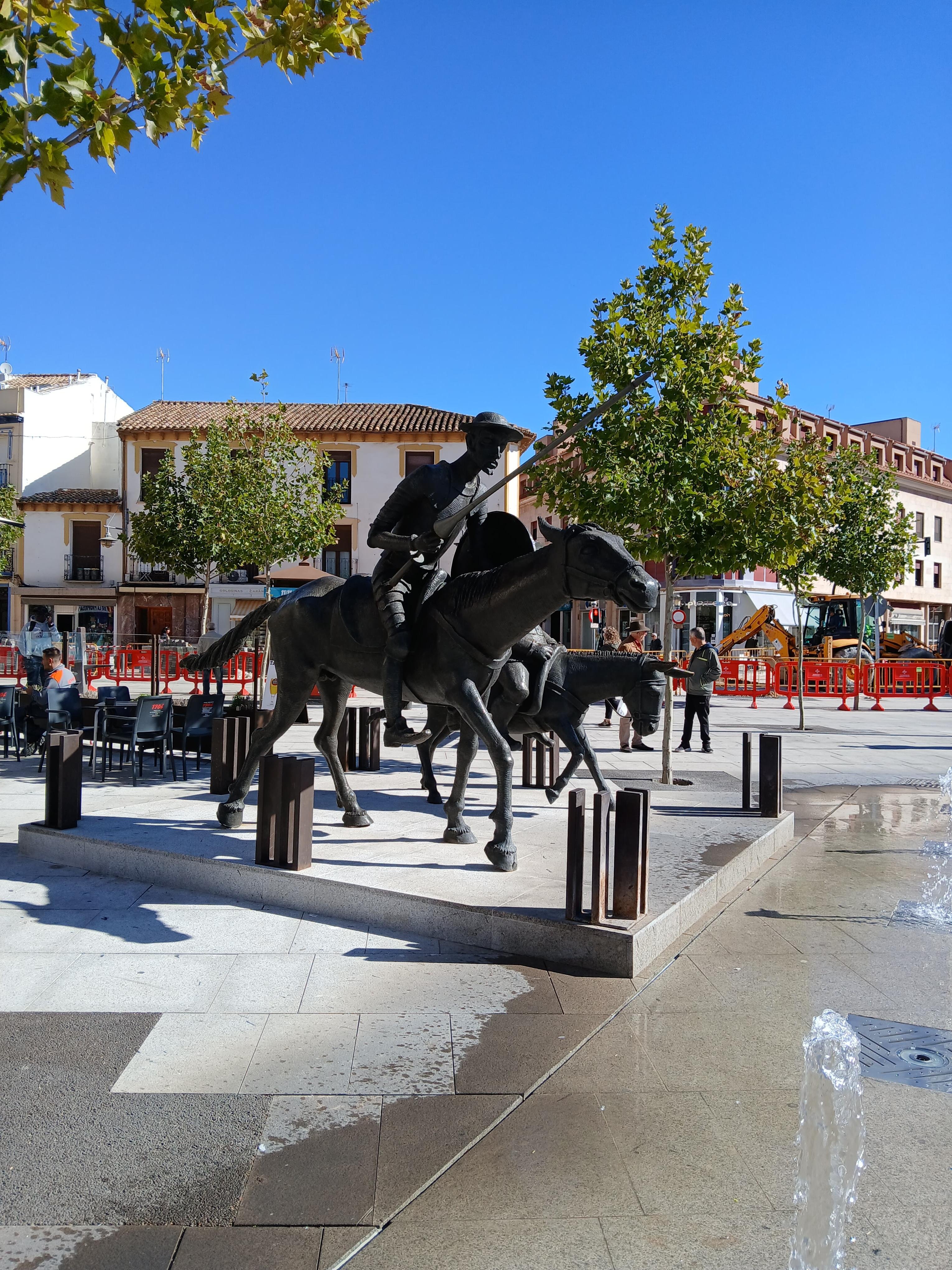 The image shows a statue of a figure on horseback, likely depicting a historical or fictional character, set in a public square. There are trees and buildings in the background, and some outdoor seating is visible to the left of the statue. The scene appears to be taken on a clear day with a blue sky.