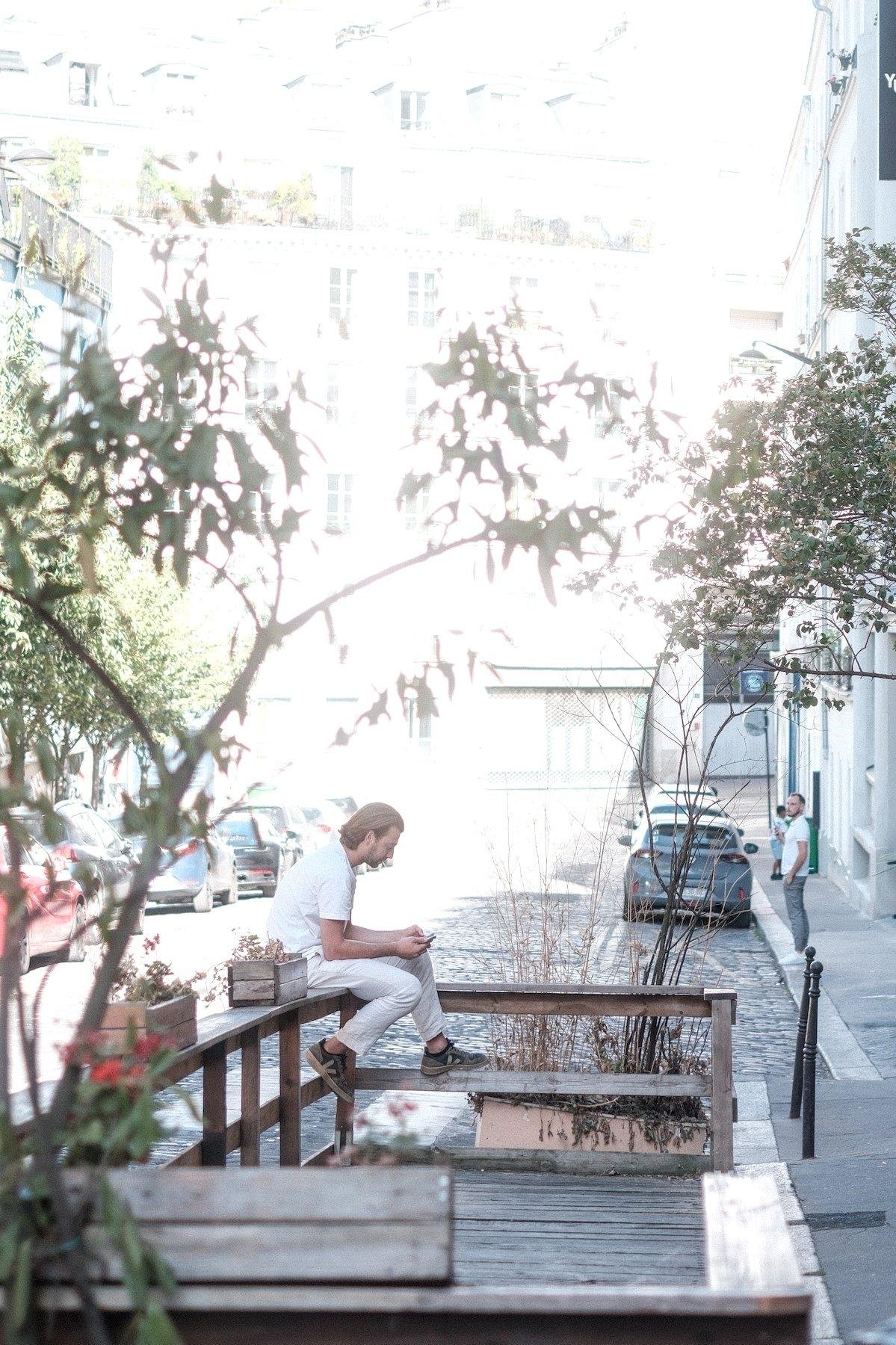 Photo verticale. Sur une terrasse en bois installée sur la chaussée, à l’ombre, un homme au cheveux mi-long, tout de blanc vêtu sauf les baskets noire, est assis sur la rambarde est consulte son téléphone. On devine une grande chaleur avec la lumière aveuglante sur les 2/3 haut de l’image