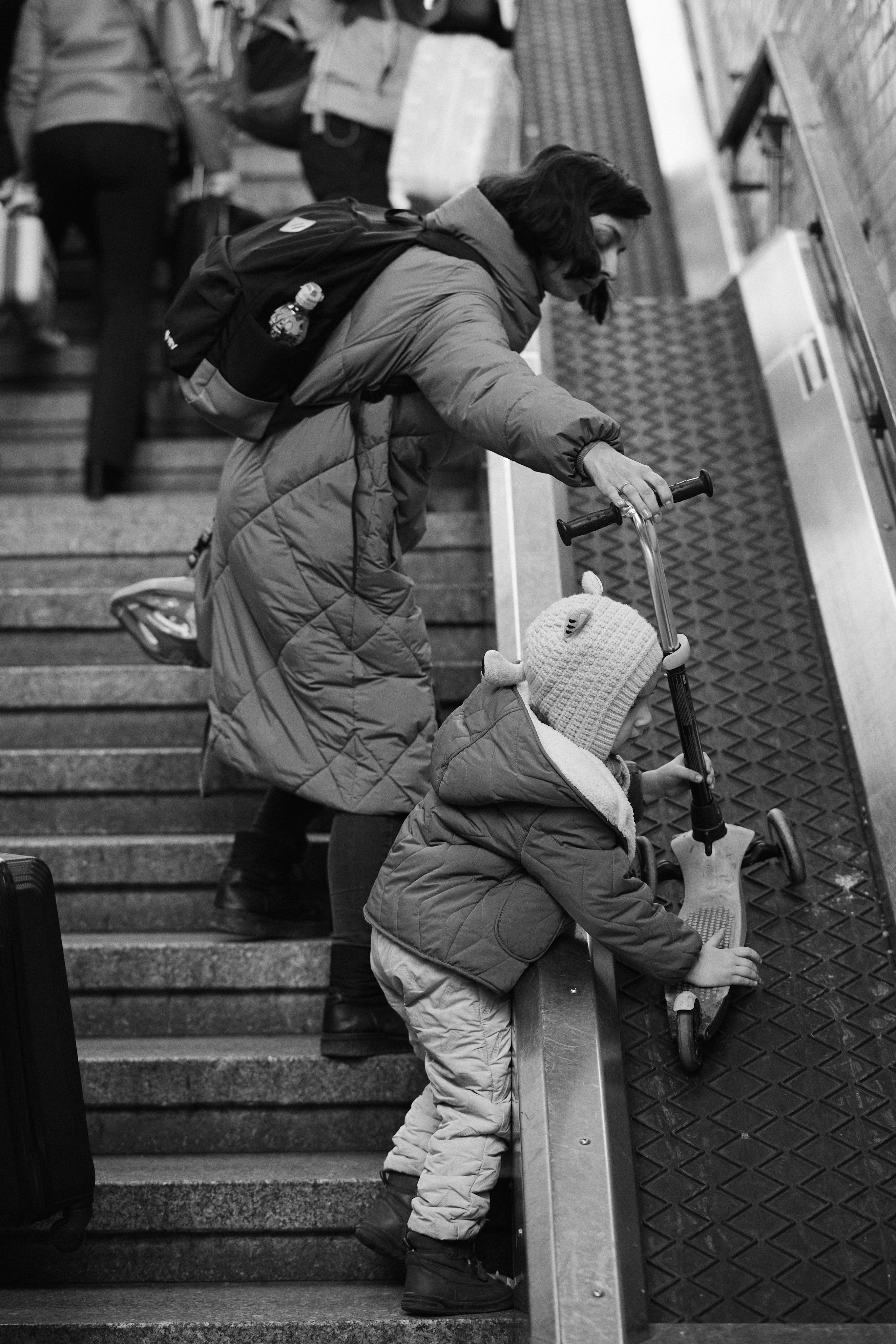 The image depicts a woman and a child on a staircase. The woman is holding a scooter as she assists the child in moving it up on a side ramp designed for wheels. Other people with luggage can be seen in the background, indicating it might be a public transit or travel location. The scene is captured in black and white.