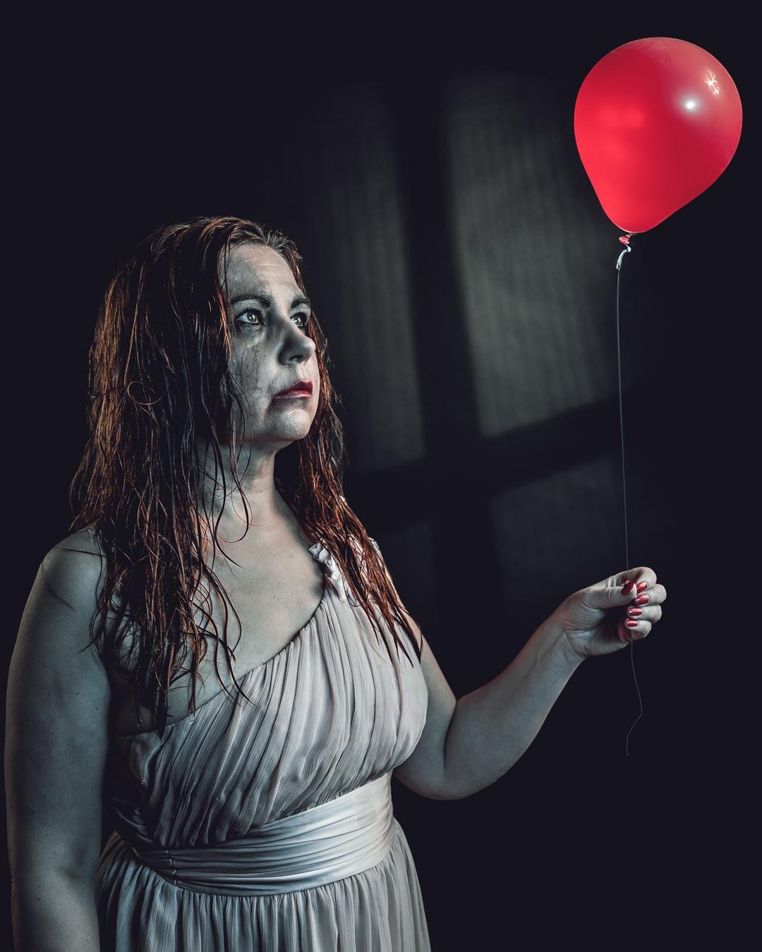 The image shows a person with a pale face holding a red balloon. They have long, wet hair and are wearing a light-colored, sleeveless dress. The background is dark and the lighting casts dramatic shadows. The overall mood appears to be eerie or dramatic.