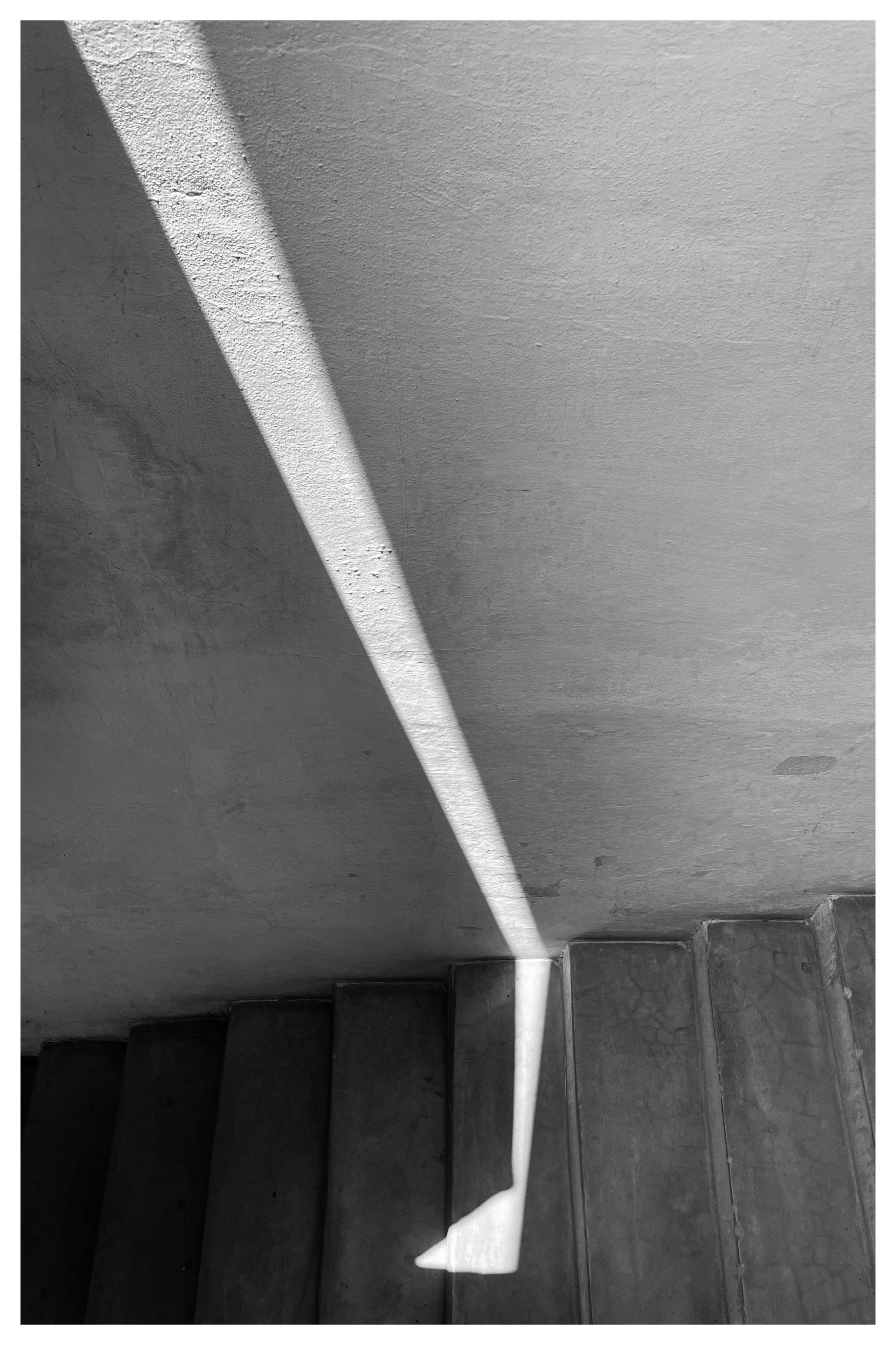 Diagonal beam of light casting a long triangular shadow across a textured wall and descending onto concrete steps below.
