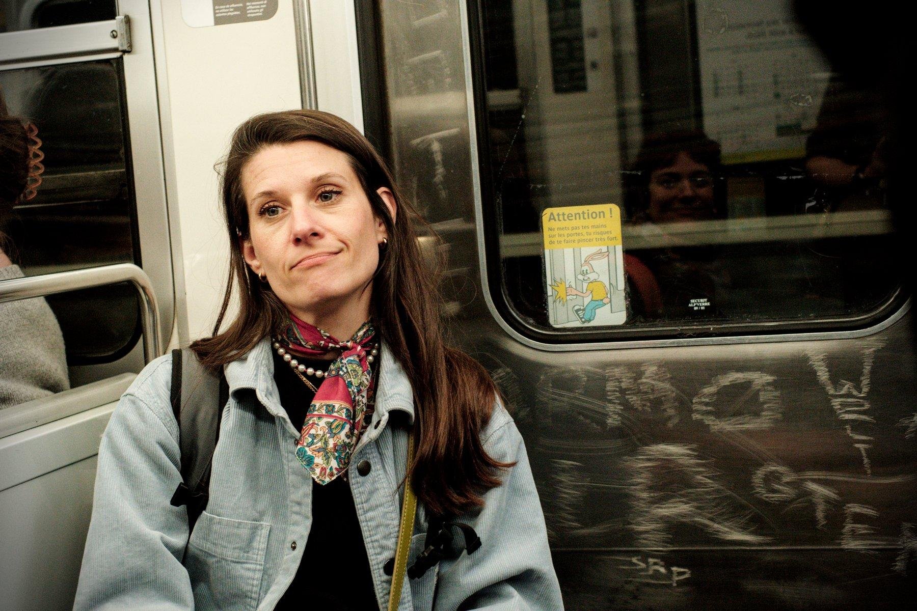 Photo horizontale. Une femme brune aux cheveux long, veste velour couleur denim, foulard coloré rouge et motif floraux, collier en perles, et d’autres chaînettes dorées, assise sur les strapontins dans le métro. Elle a le visage exprimant la compréhension, avec les lèvres mi pincées, mi sourire.