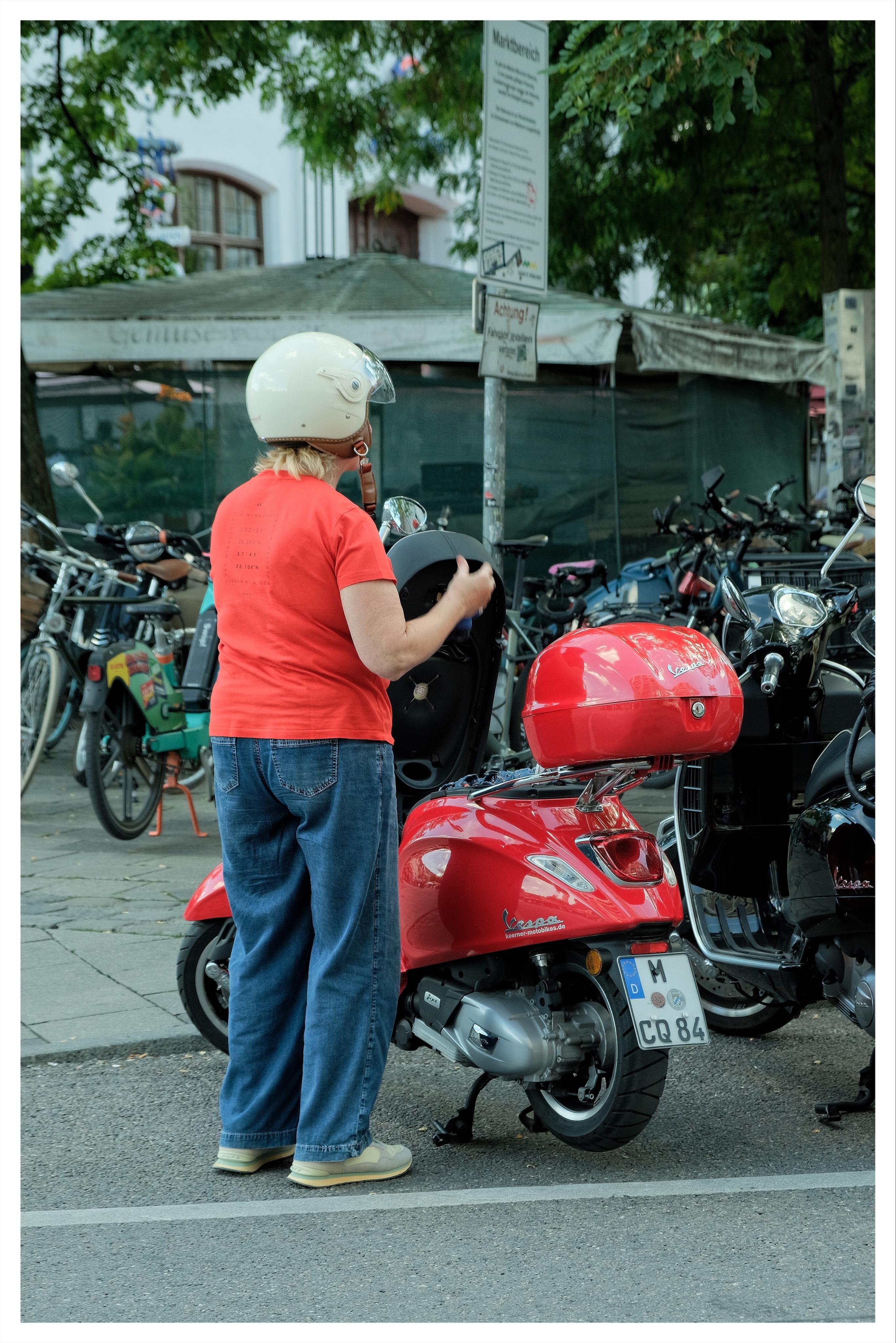 Person in a white helmet, red T-shirt, and jeans stands beside a red Vespa-style scooter with a rear cargo box, parked at the curb near a cluster of bicycles and motorcycles; a street sign and leafy trees are in the background.