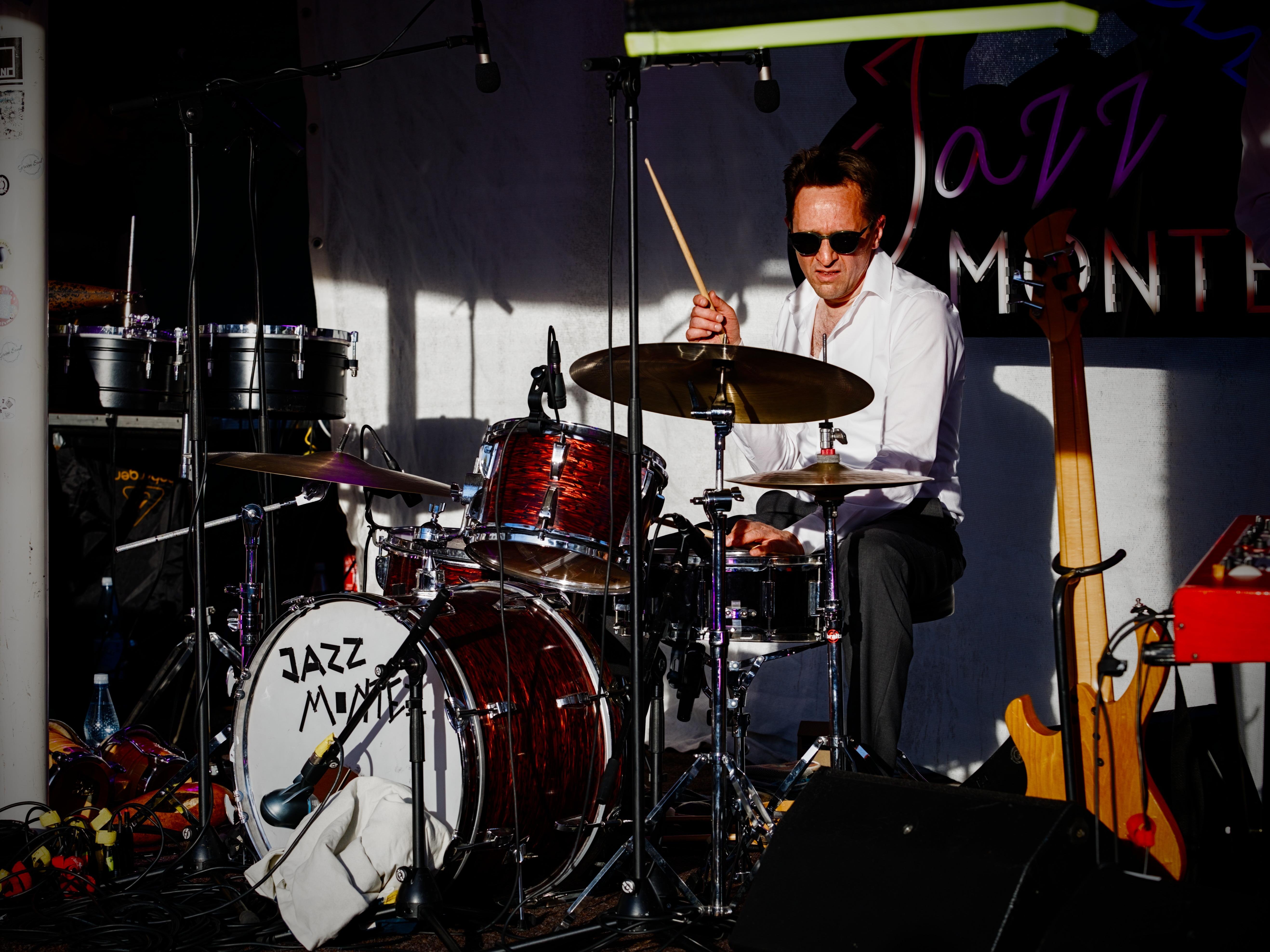 A man playing the drums at a festival. The back of kick drum reads "Jazz Montez", the name of the festival. The logo also partially appears behind him. He is bathed in sunlight with harsh shadows everywhere on stage. He plays on a typical jazz kit with a reflective dark red finish. His pose is very iconic, with one hand playing a cross-stick on the snare drum while the other one holds the stick in the air, ready to hit the crash cymbal quite hard. His focused look is strengthened by the white shirt and the sunglasses he is wearing. A wooden bass guitar is standing on the right-hand side, held by a rack.