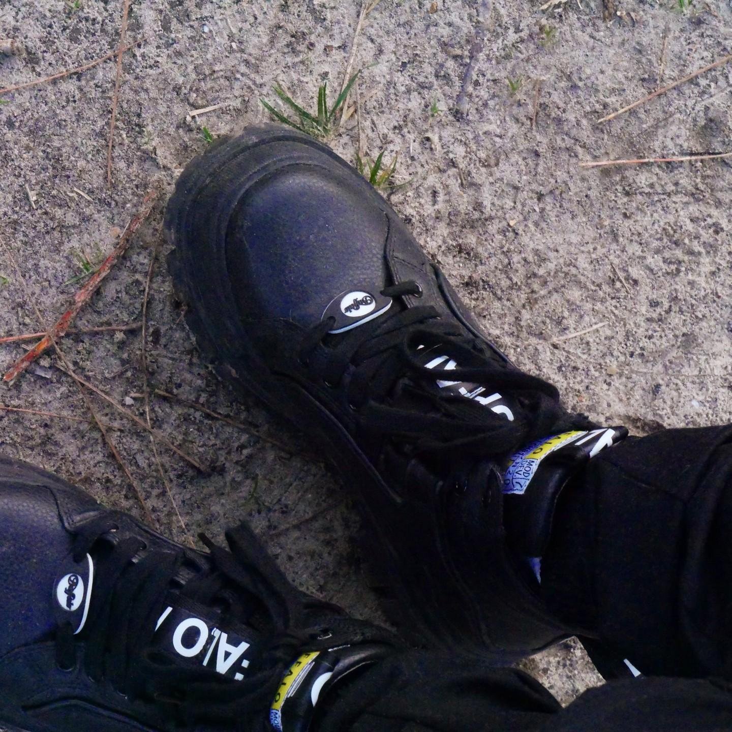 The image shows a pair of black Buffalo shoes on a sandy, outdoor surface. The shoes have thick soles and laces. There's some text and logo on the shoes.