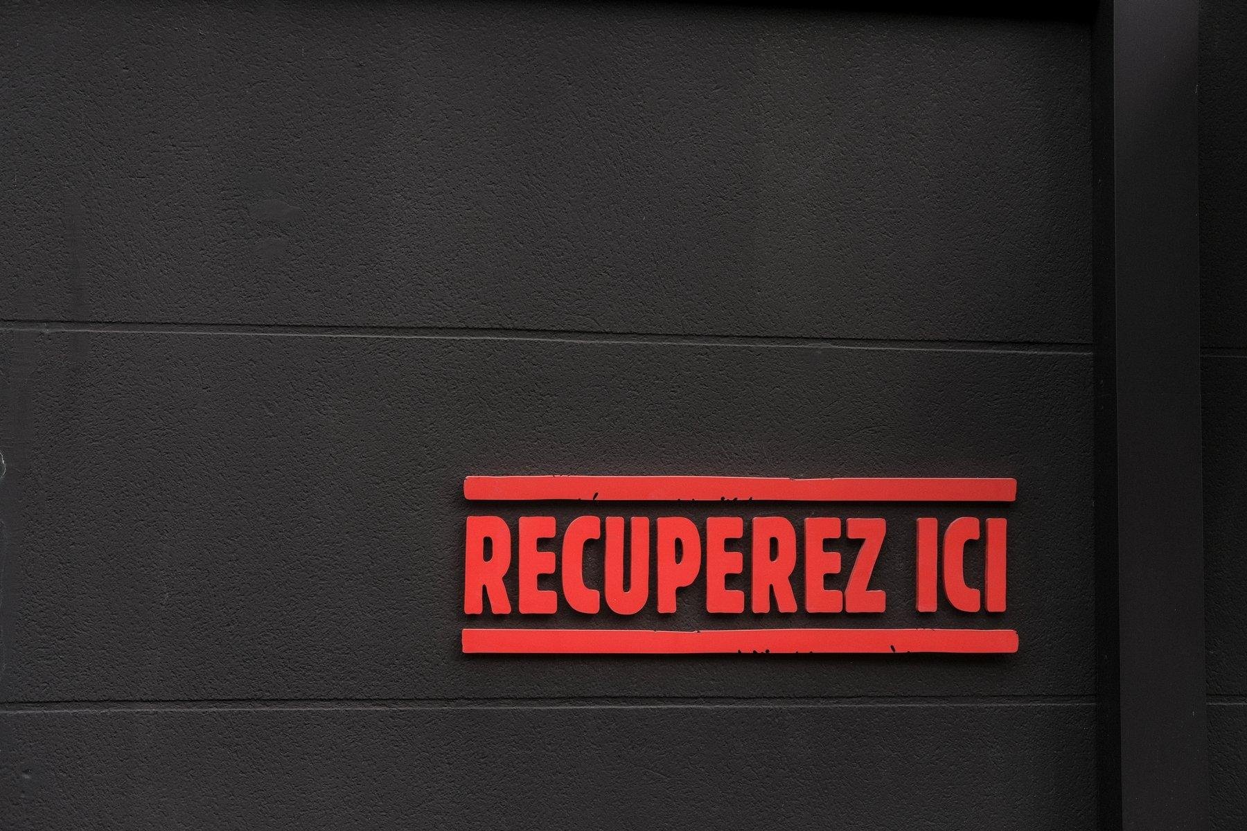 Photo horizontale. Un mur marron très sombre presque noir, et dessus, en rouge, les mots en lettres capitales «&nbsp;RECUPEREZ ICI&nbsp;» avec un trait rouge au dessus et un autre en dessous, occupant le quart inférieur droit de l’image